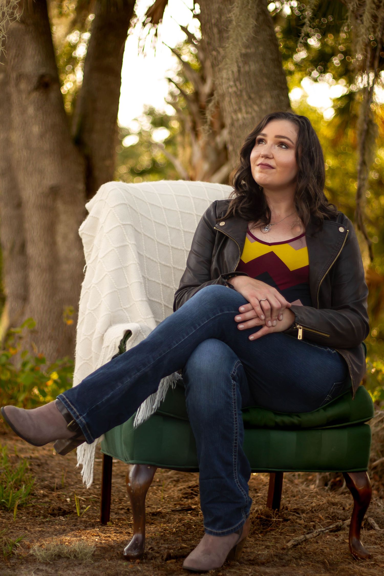 Young woman in leather jacket sitting in green velvet chair outdoors at sunset.