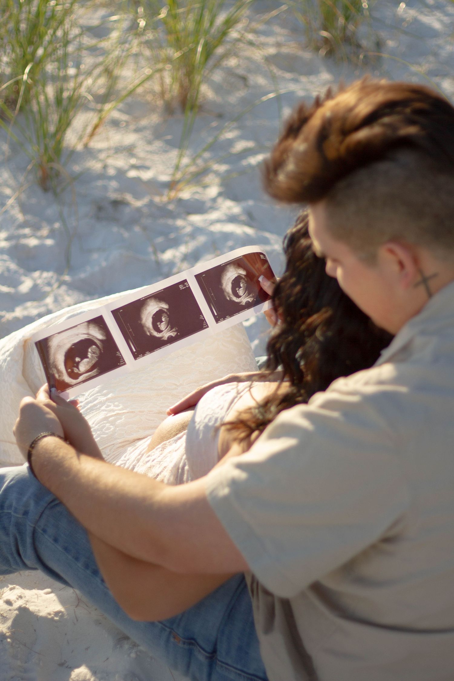 Couple sitting on sand dunes together looking at ultrasound photos in sunlight.