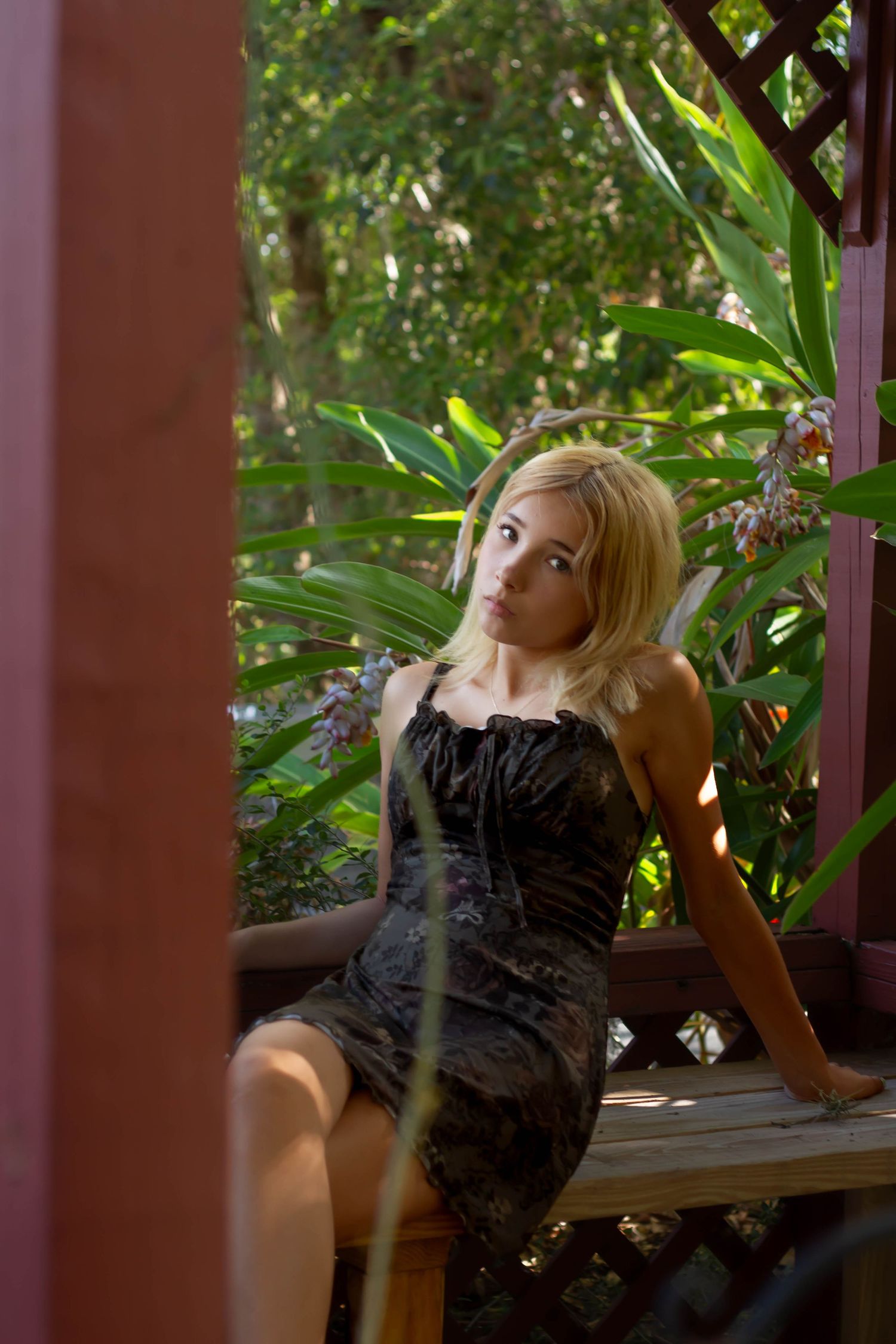 Young blonde woman in dark floral dress sitting in garden gazebo surrounded by tropical plants.