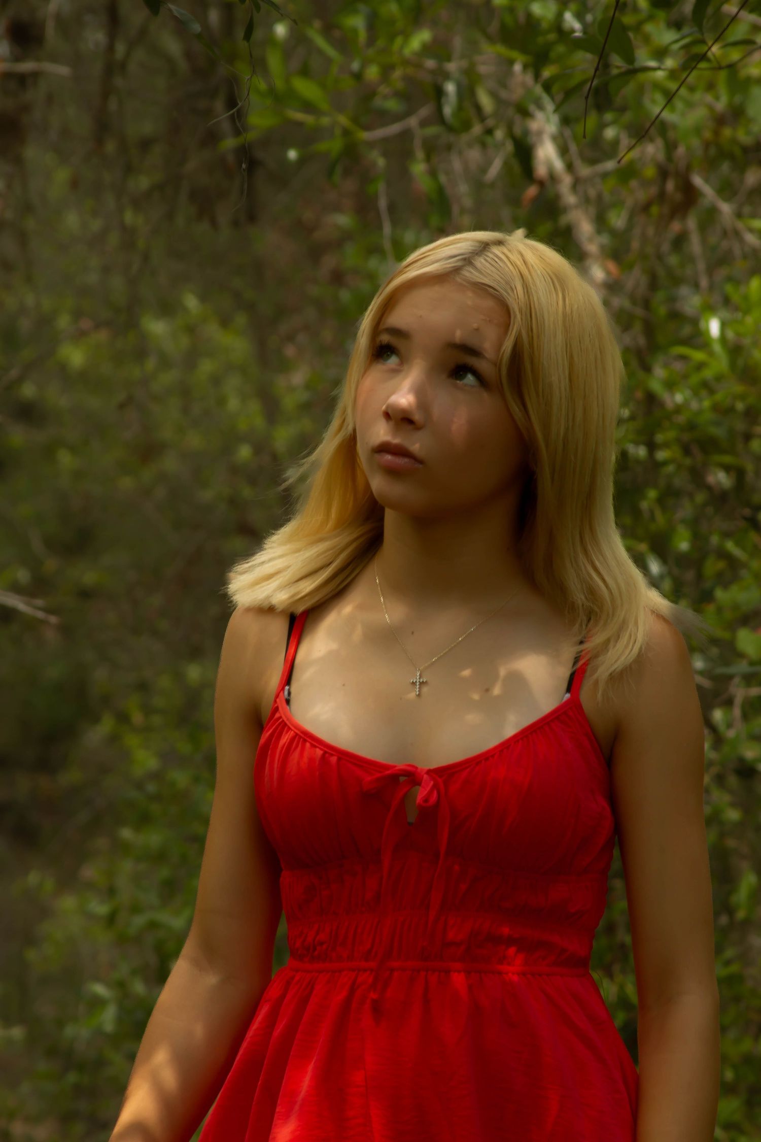 Young blonde woman in a red sundress with cross necklace gazing upward in a lush green forest.