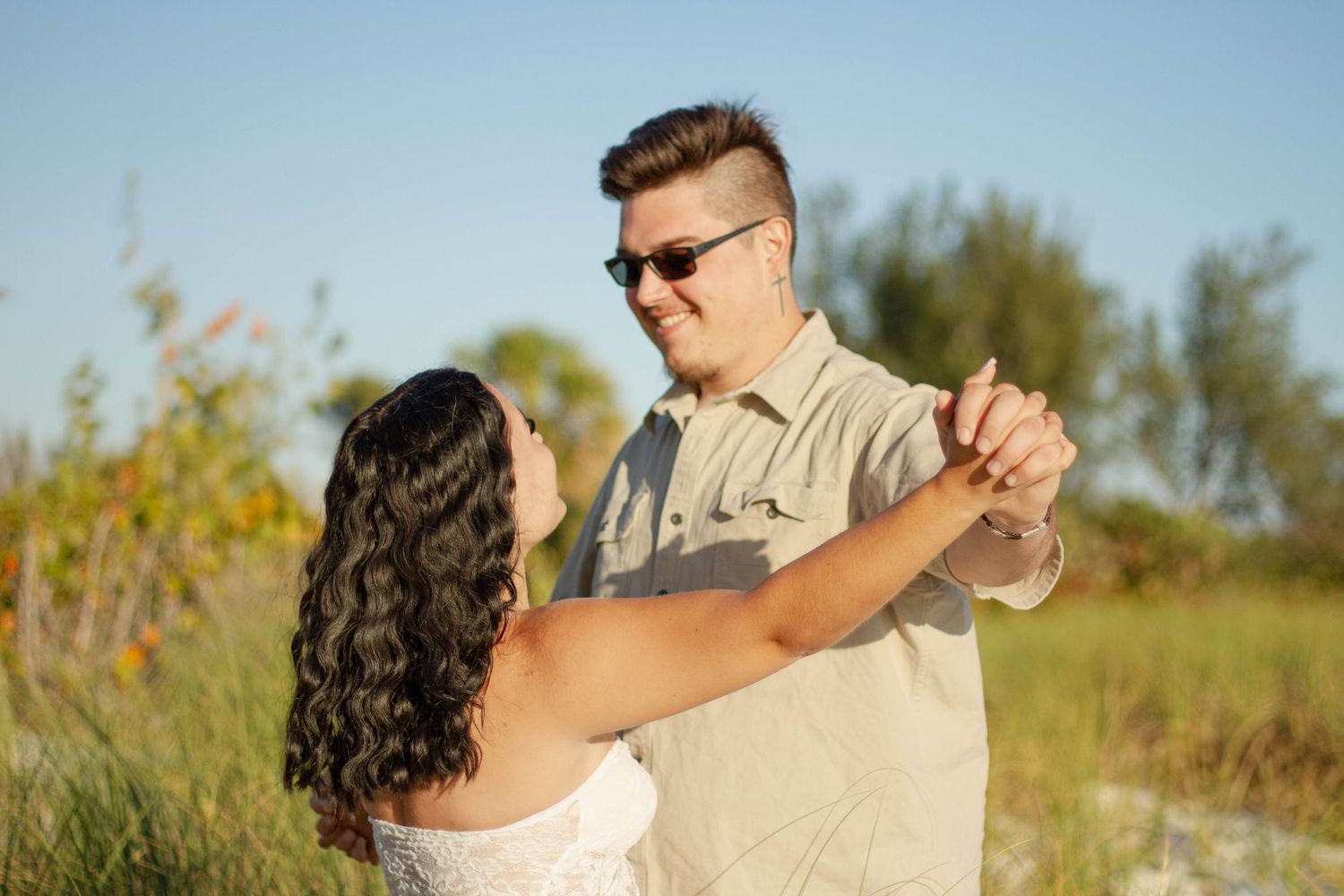 Couple dancing together in sunlit field, man in sunglasses and beige shirt, woman in white dress.