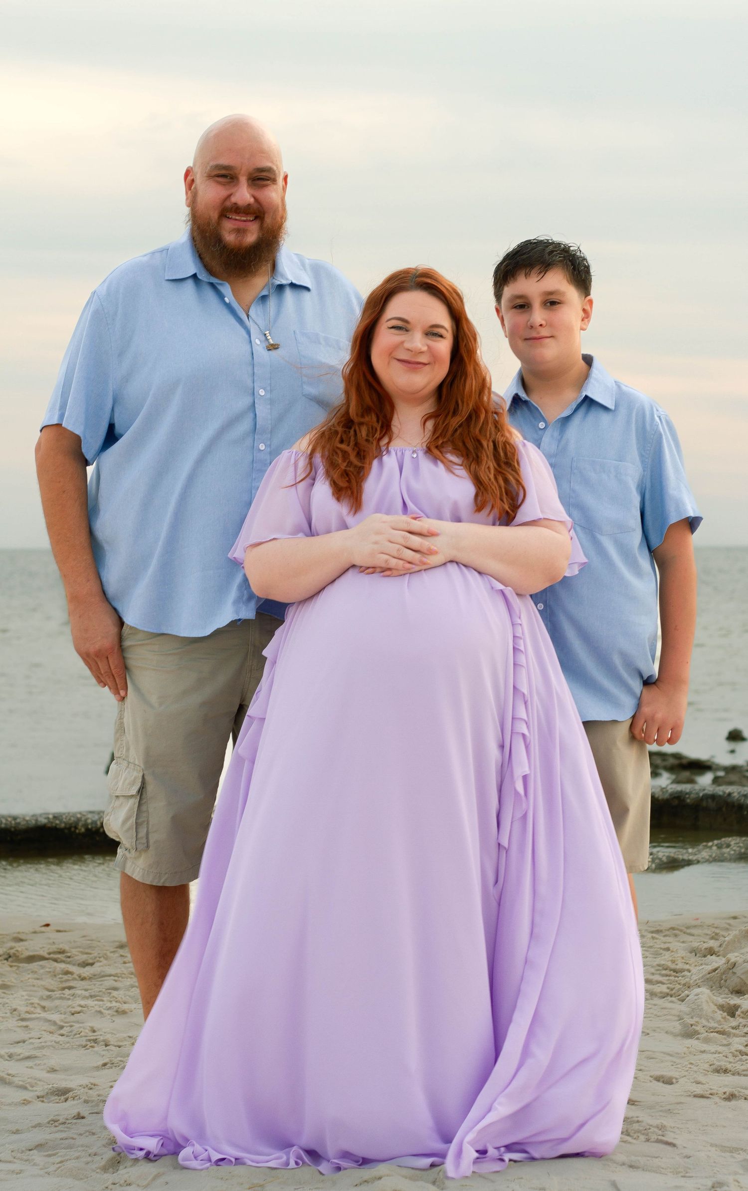Pregnant woman in lavender gown poses with bearded man and teen boy in blue shirts on beach.