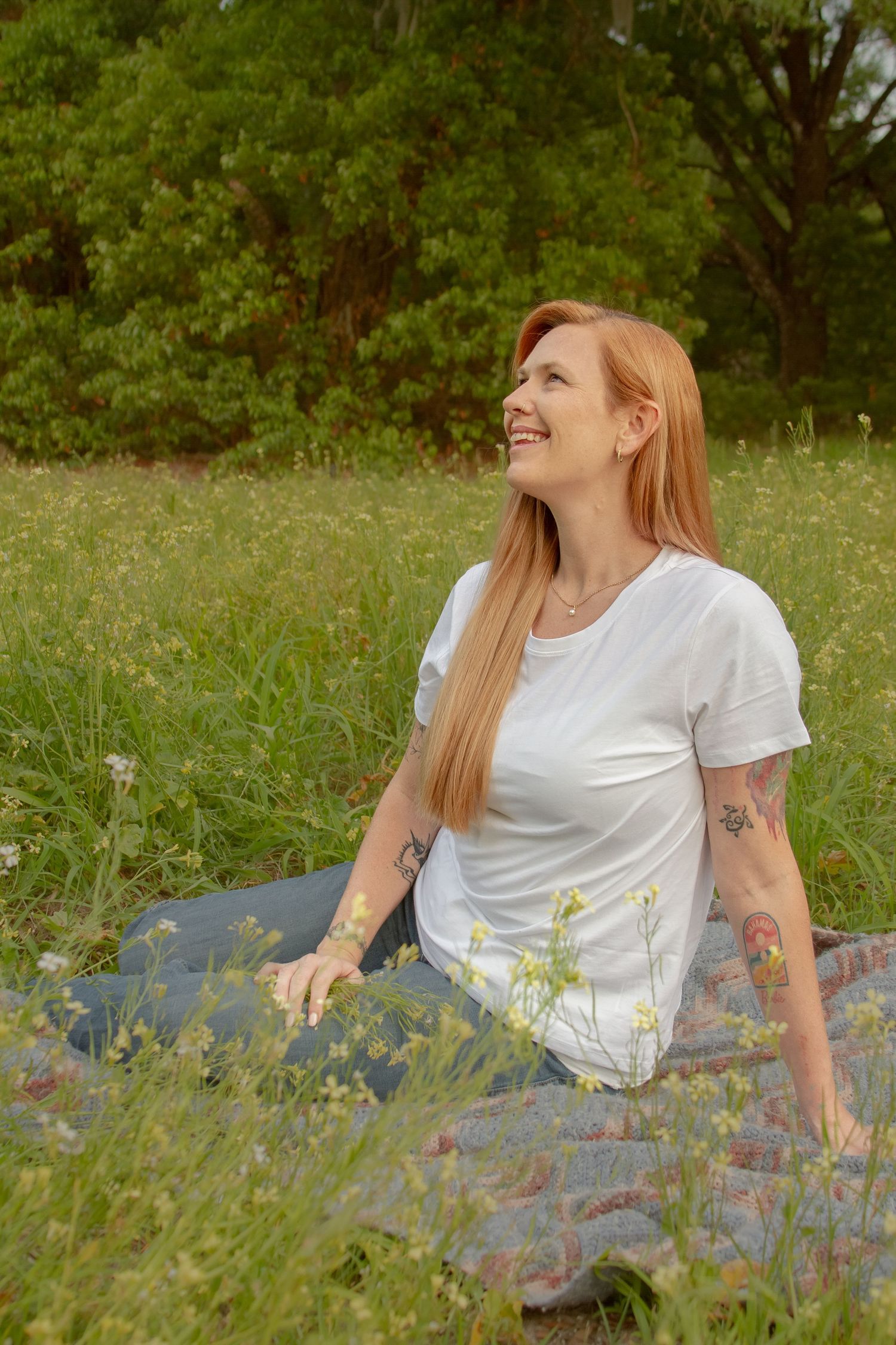 Smiling redhead woman sitting on blanket in wildflower meadow wearing white t-shirt with tattoos visible on arms.