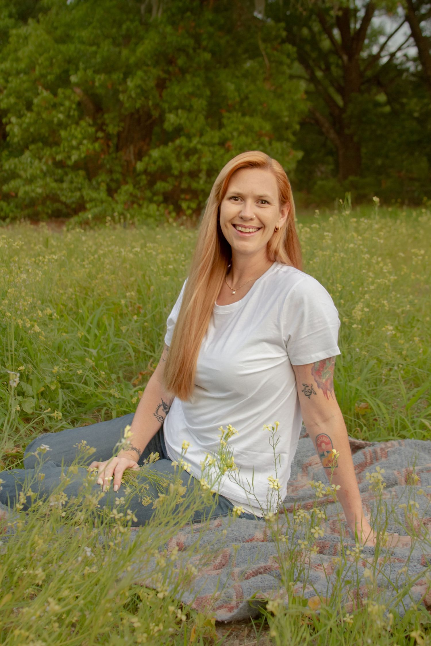 Smiling redhead woman with tattoos in white tee sitting on blanket in lush green meadow.