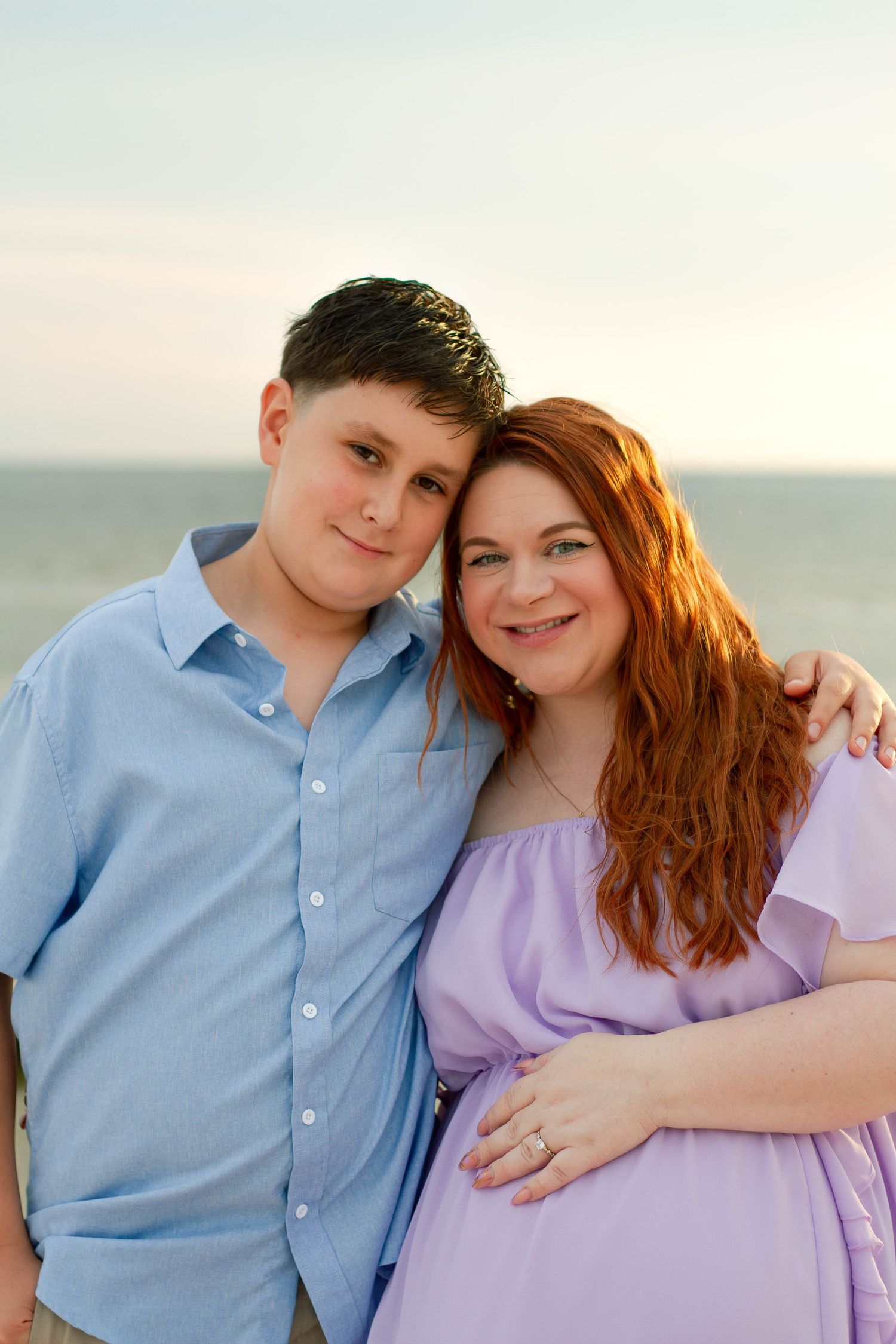 Smiling pregnant redheaded woman and young boy pose together on the beach at golden hour.