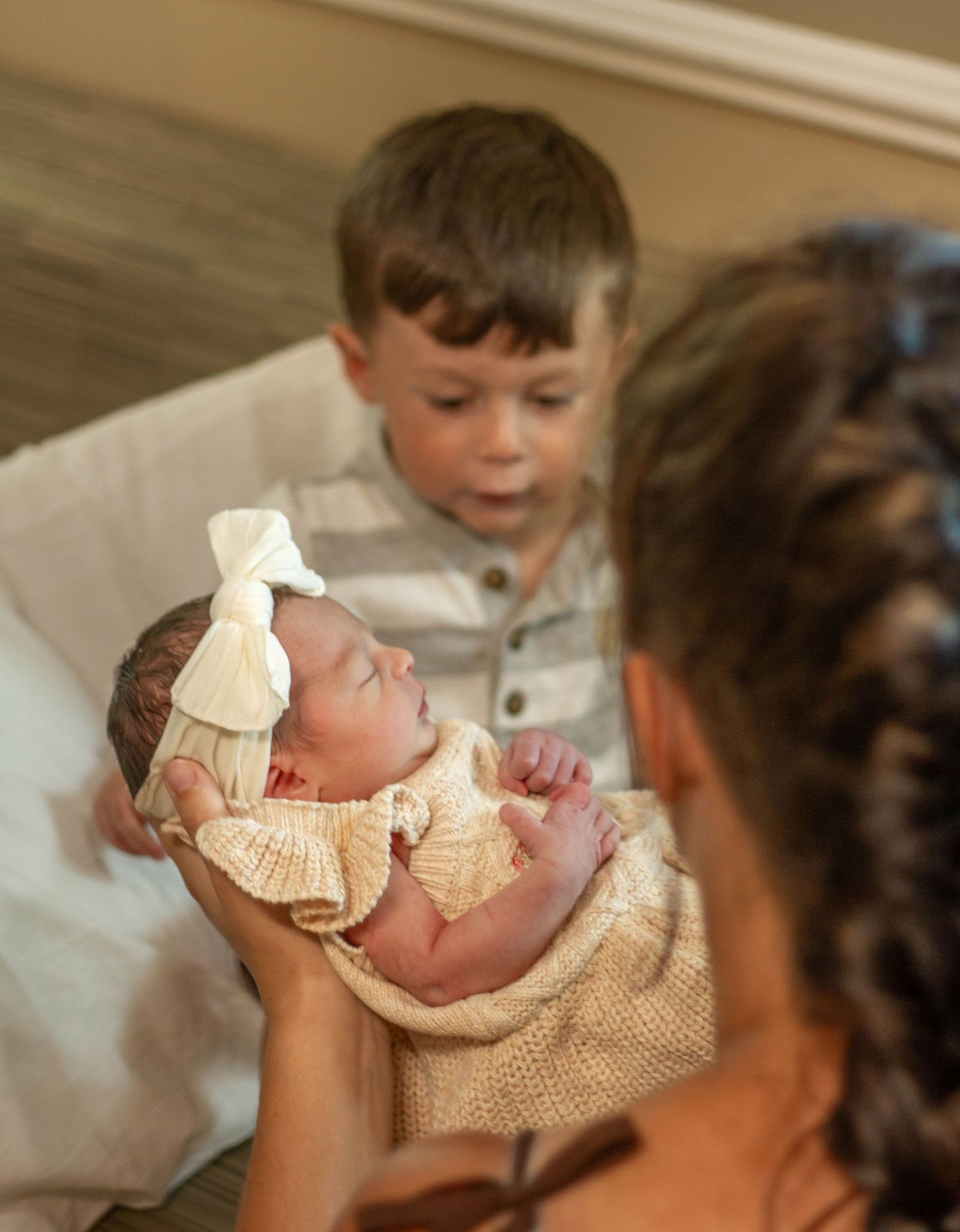 A mother holds her newborn baby girl in a white bow while an older brother gazes at her lovingly.