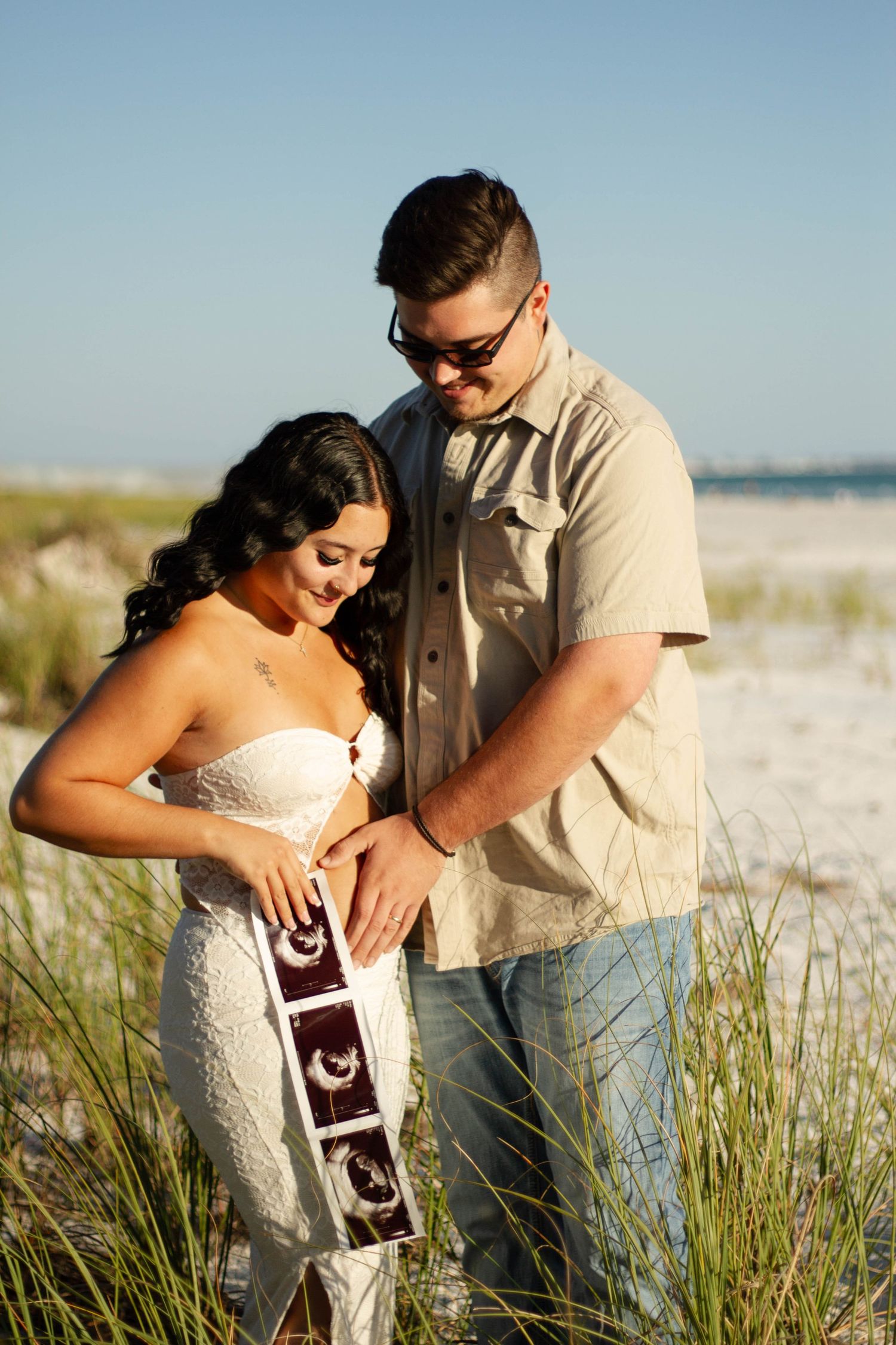 Couple smiling at ultrasound photos on sandy beach, celebrating pregnancy together.