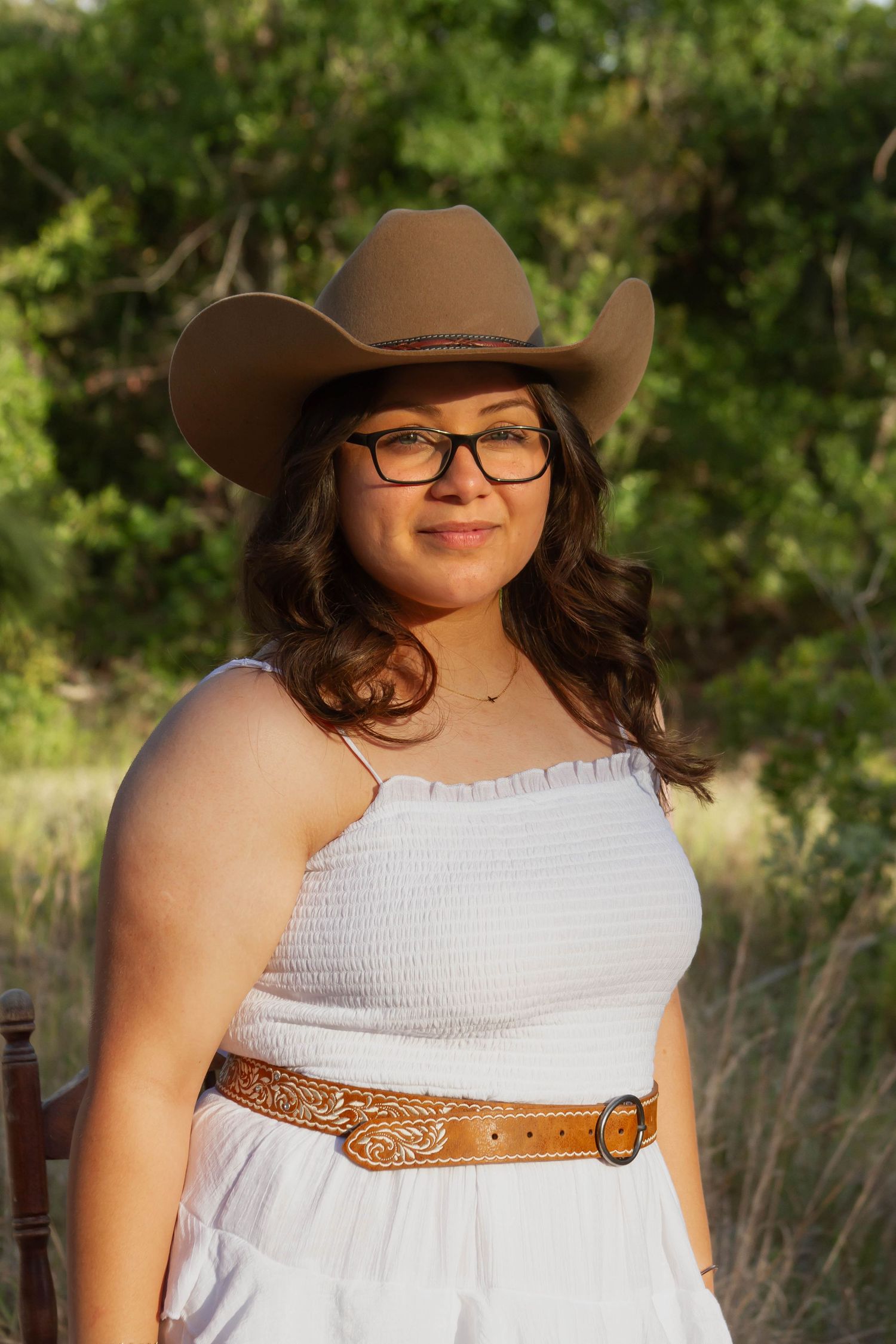 Young woman in cowboy hat and glasses wearing white dress with brown belt outdoors.