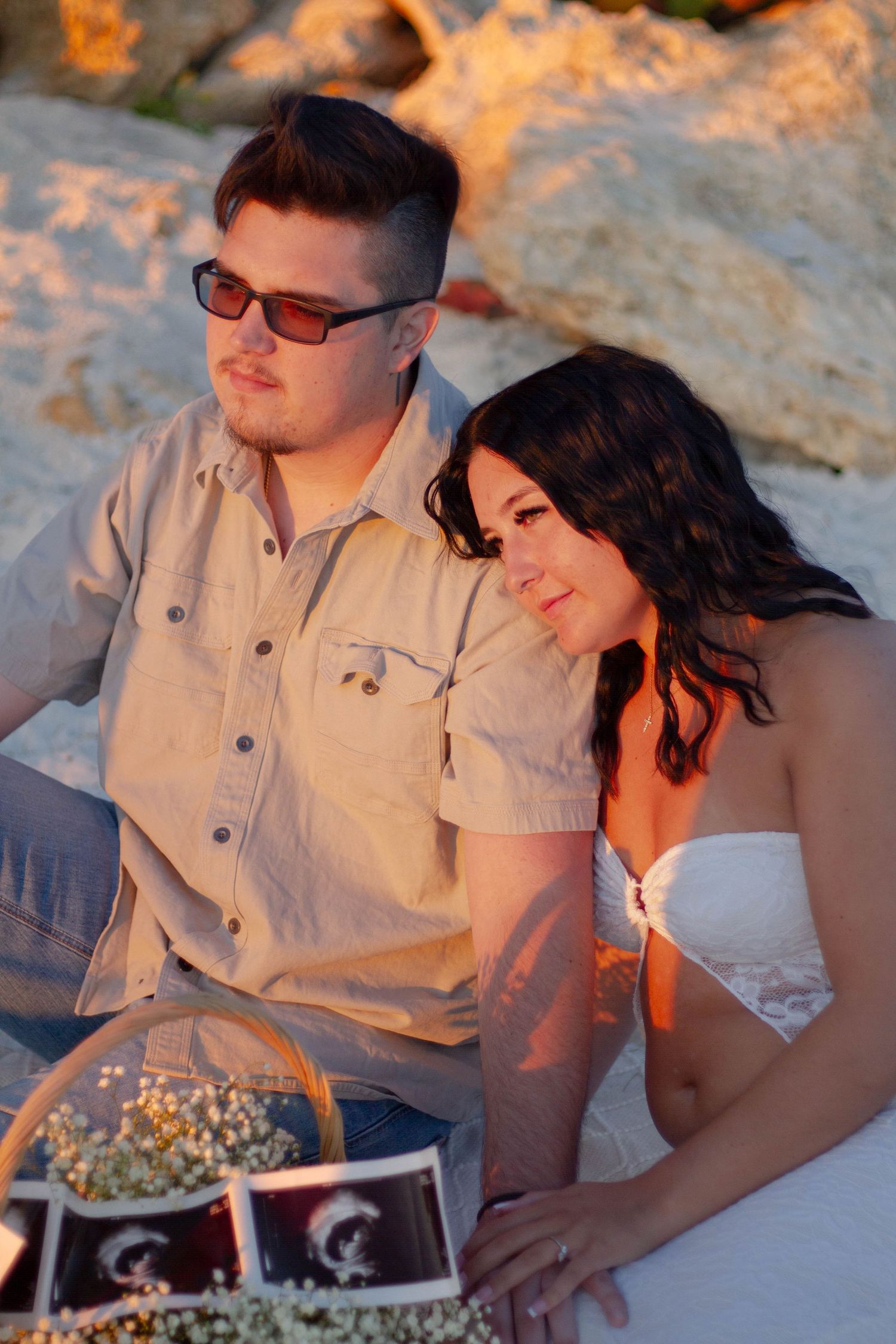 Expecting couple sitting together on beach at golden sunset with ultrasound photos and flower basket nearby.