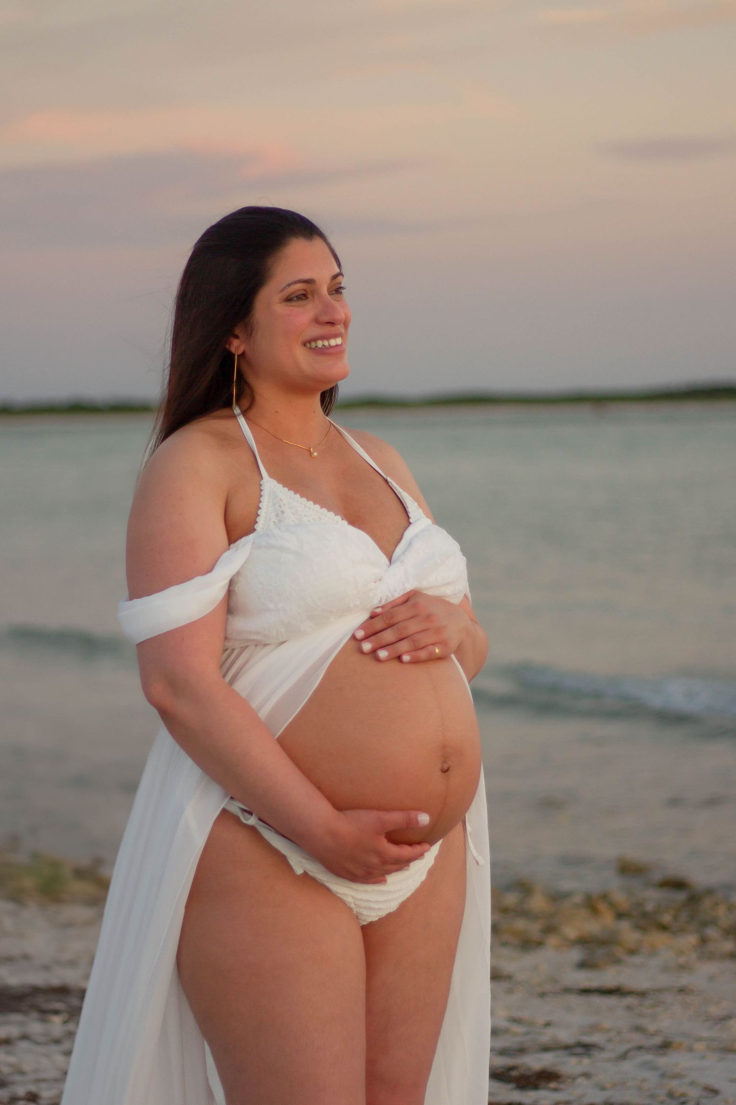Smiling pregnant woman in white outfit and sheer wrap cradles her bare belly on a beach at sunset.