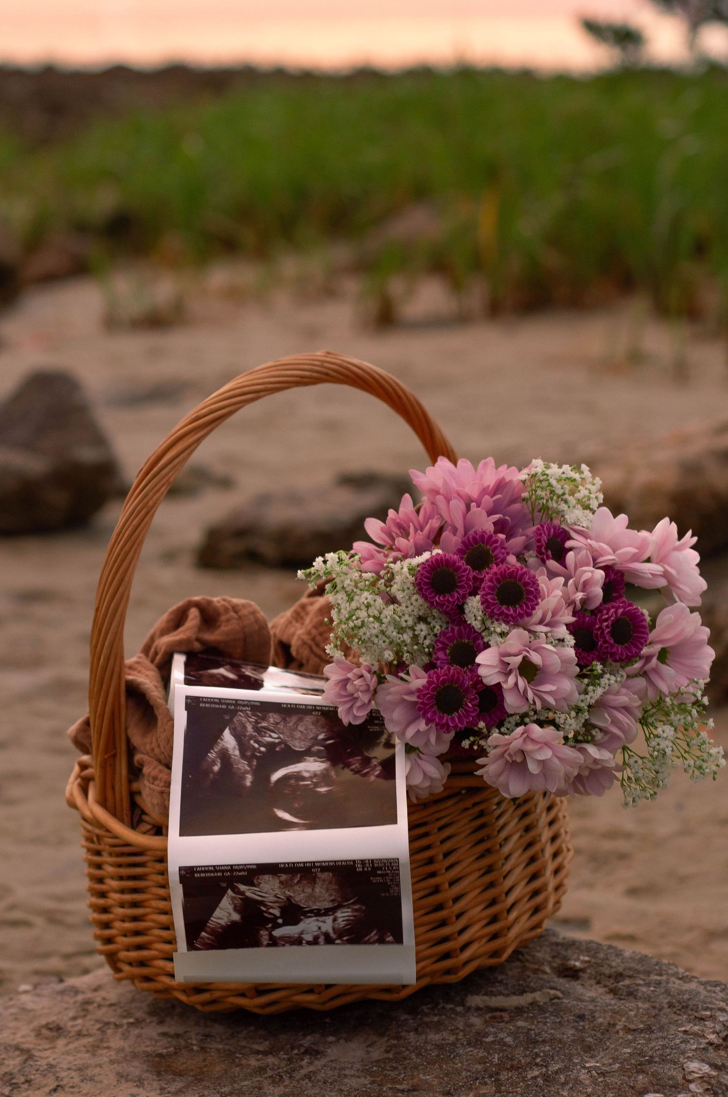 Wicker basket with purple flowers, ultrasound photos, and baby items on sandy beach at sunset.