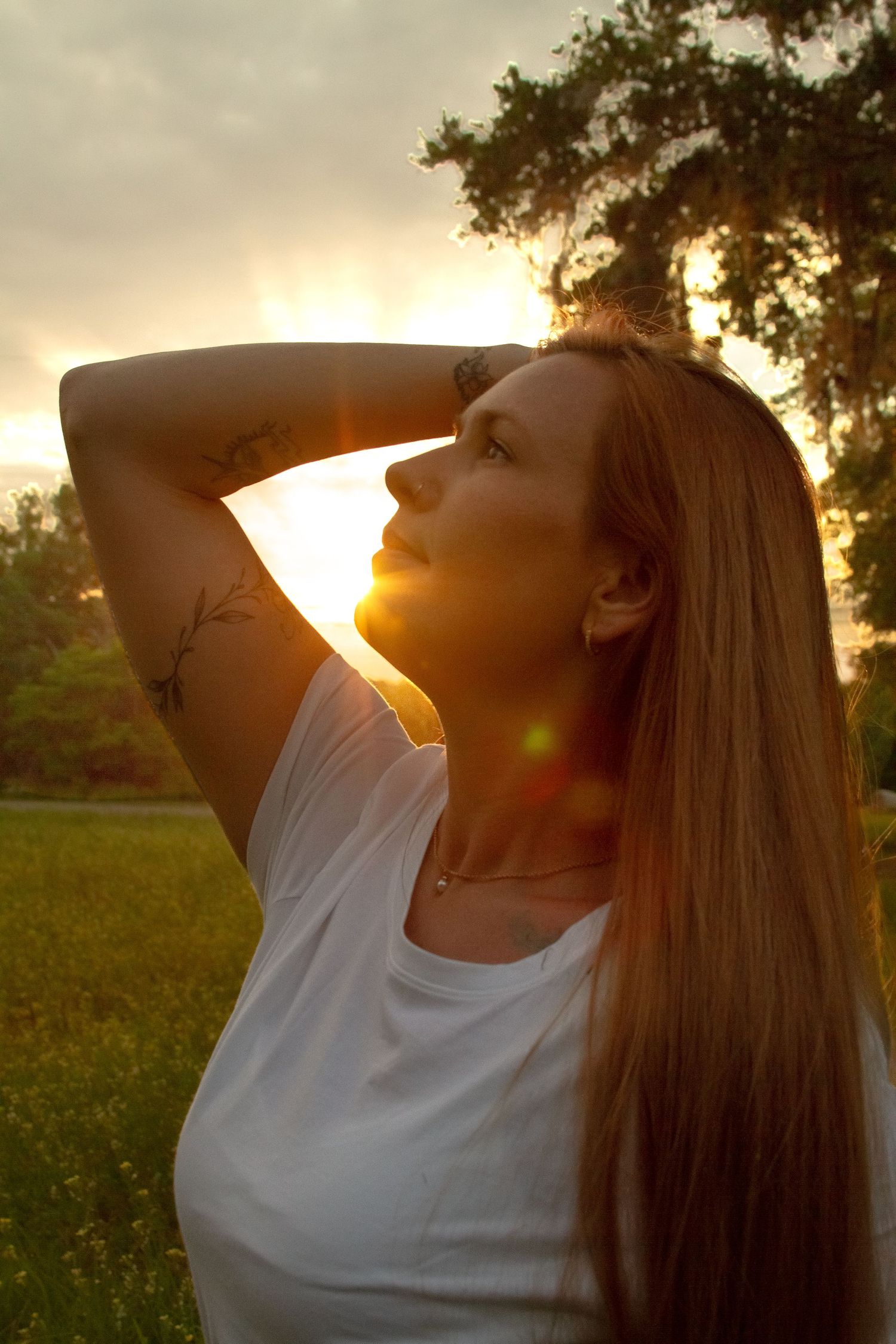 Redhead woman in white t-shirt with arm tattoo gazing upward during golden sunset in green meadow.