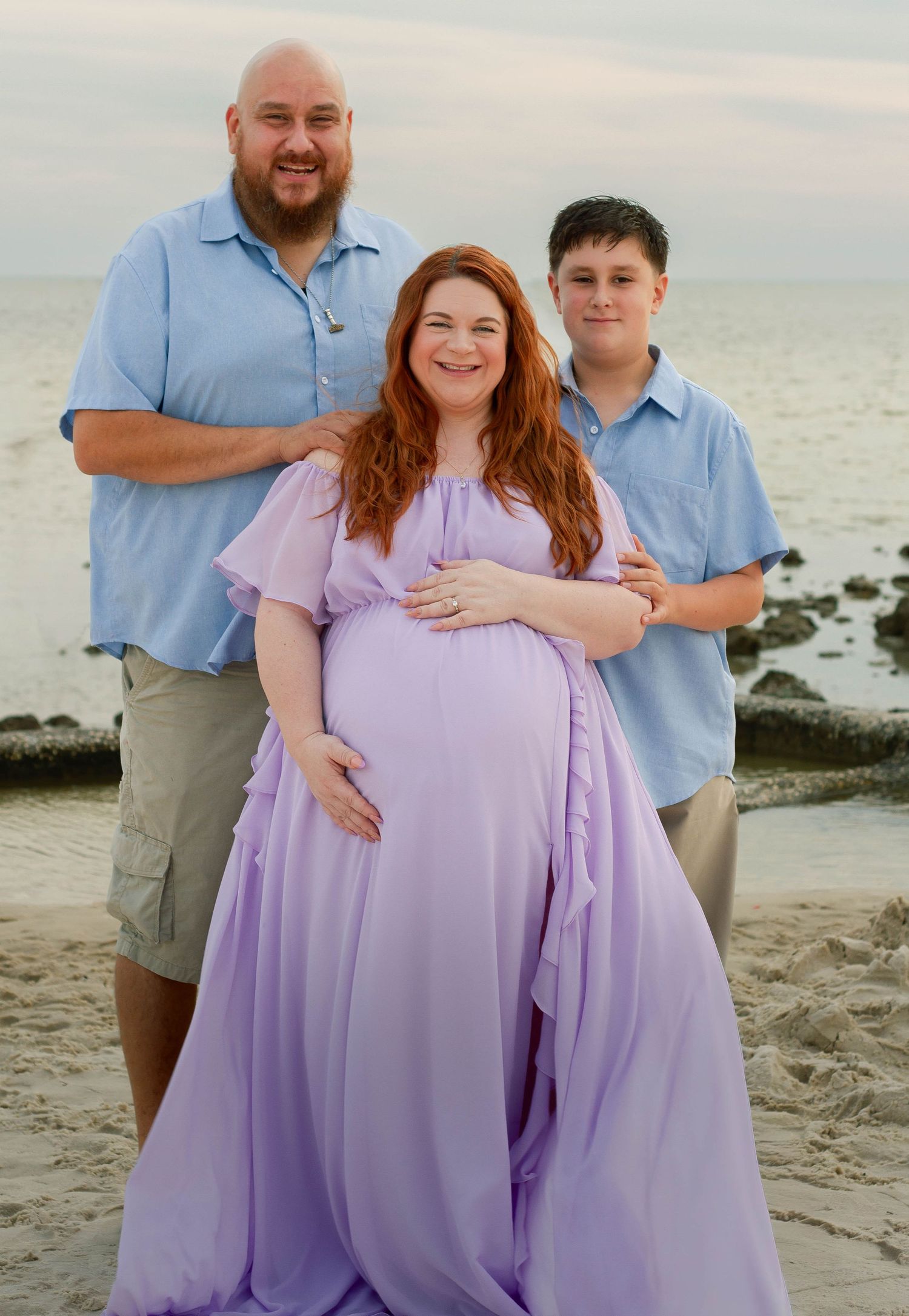 Pregnant woman in flowing lavender gown poses with husband and teenage son on sandy beach.