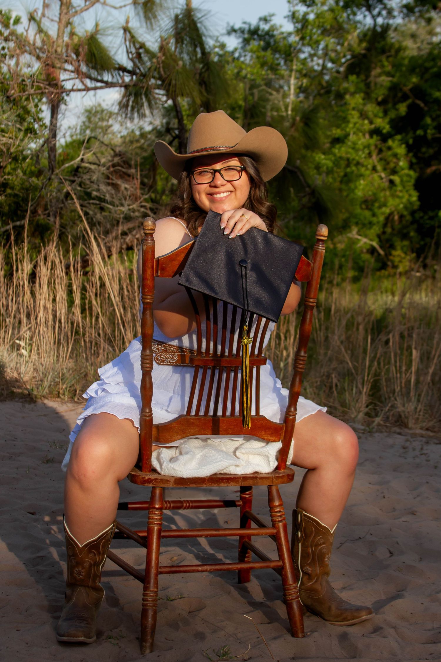 Young woman in cowboy hat holding graduation cap while sitting on wooden chair outdoors.