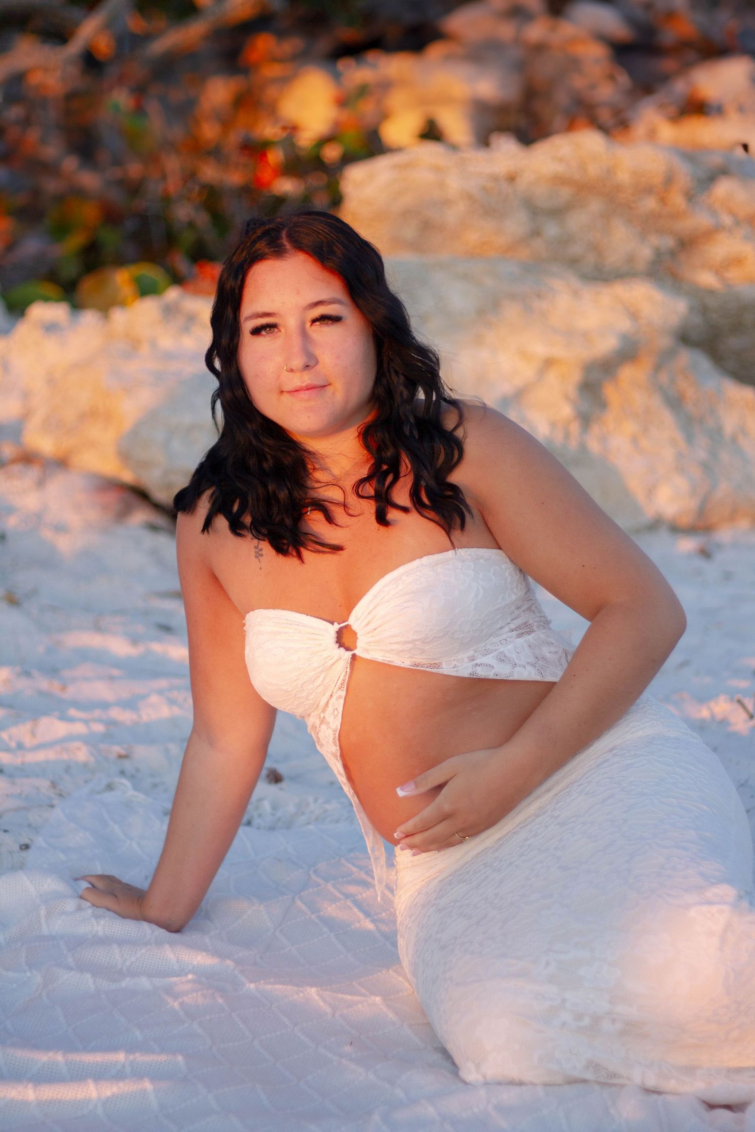 Pregnant woman in white strapless dress sitting on white sand beach at golden hour.