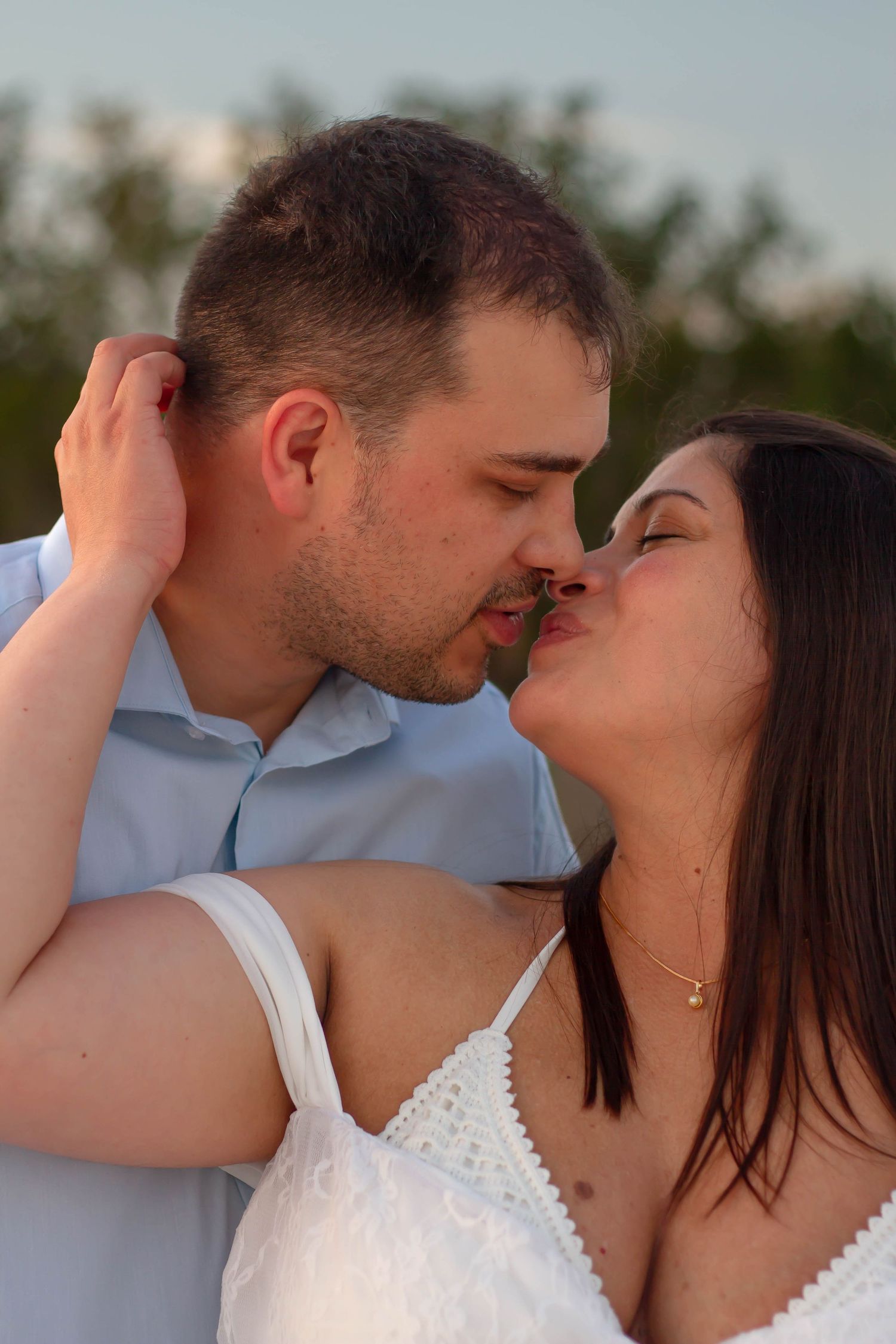 Couple shares a romantic kiss outdoors with lush green trees visible in the background.