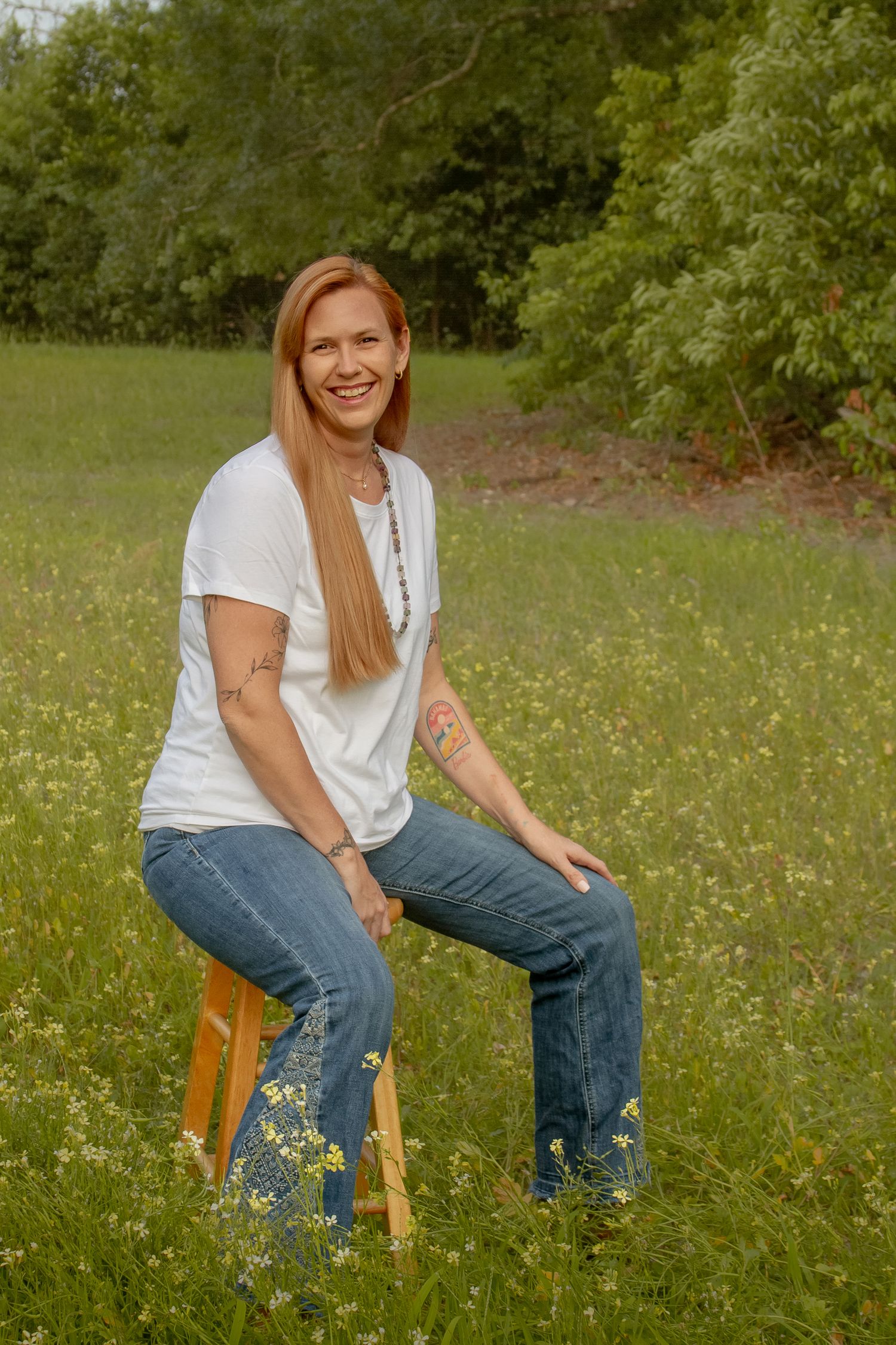 Laughing redhead woman in white tee and jeans seated on stool in green wildflower field.