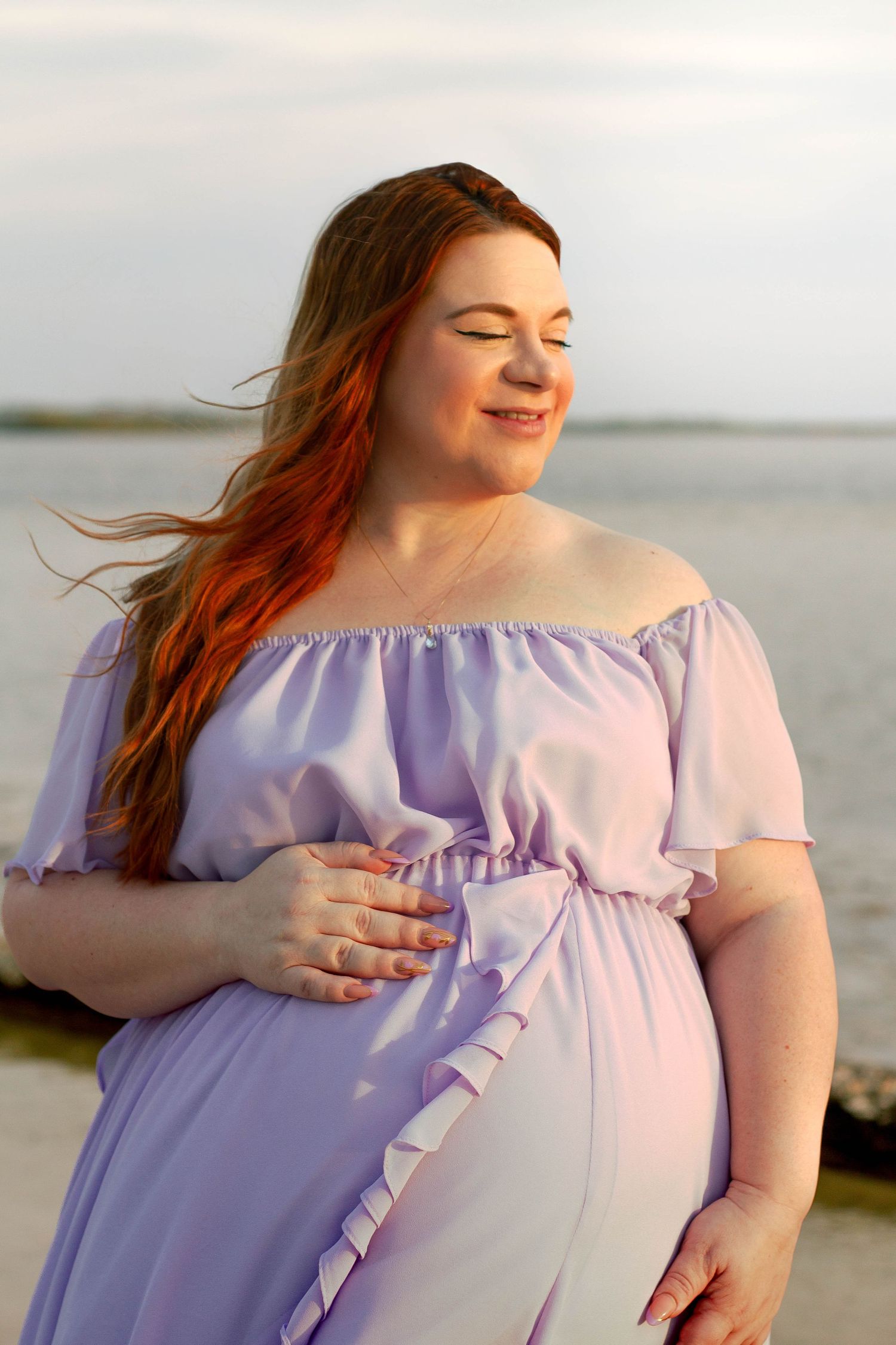 Pregnant redheaded woman in lavender off-shoulder dress cradles her baby bump on a breezy beach.