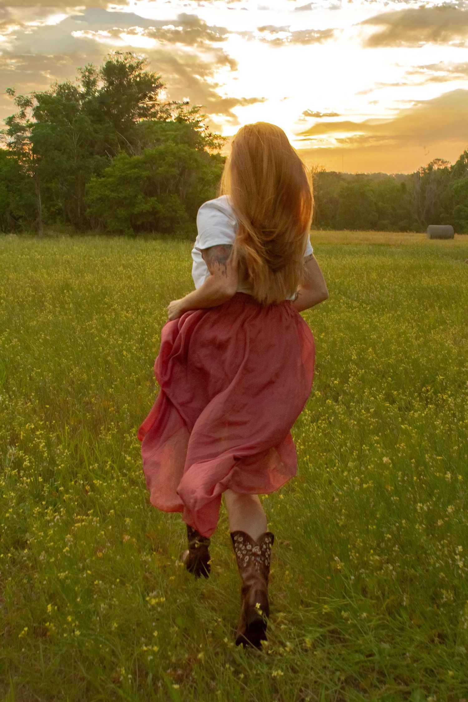 Redhead woman in white top, red maxi skirt and cowboy boots running through a golden sunset meadow.