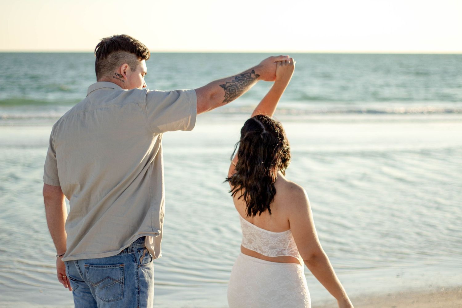 Couple holding hands on beach at sunset, man spinning woman in white dress.