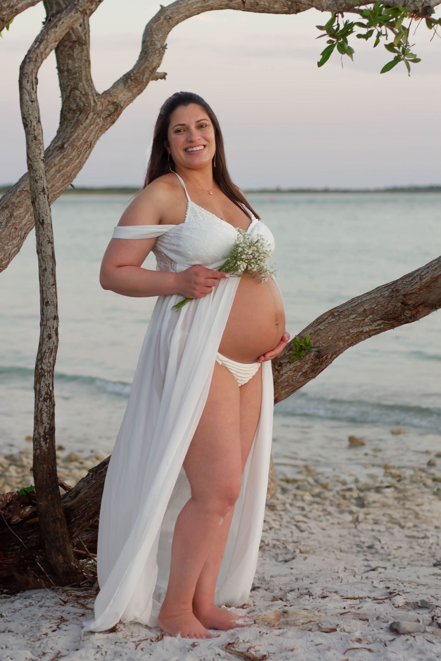 Smiling pregnant woman in a white sheer wrap holding flowers, posing gracefully by a beachside tree.