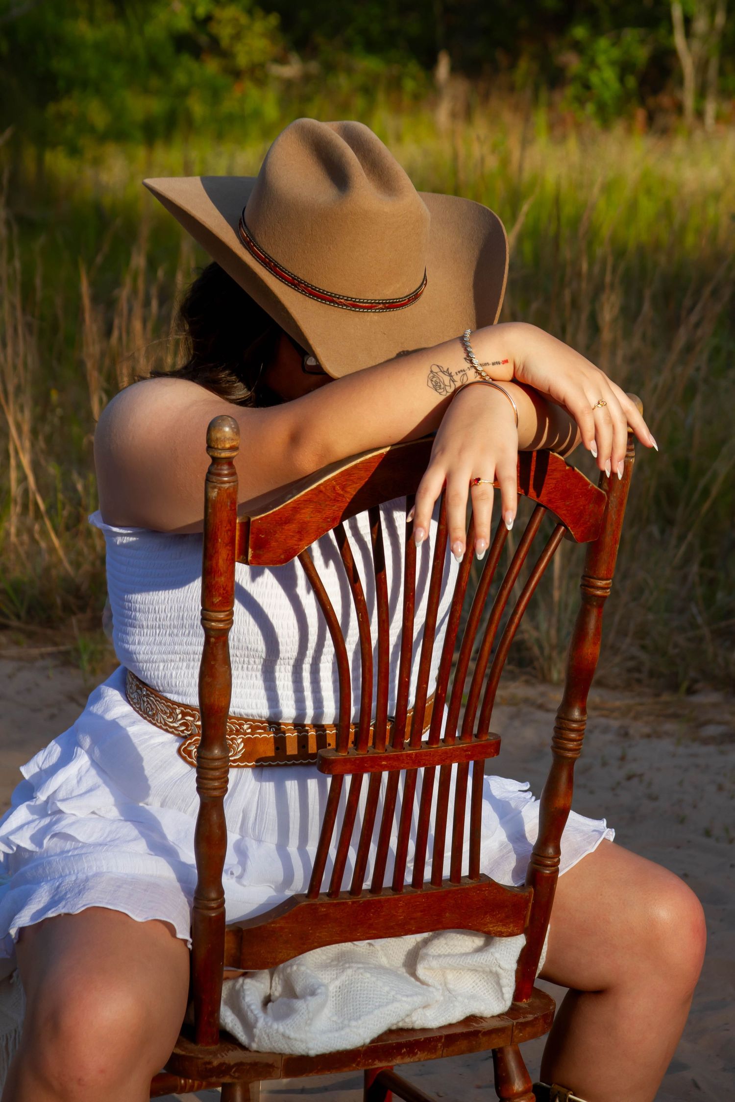 Young woman in cowboy hat and white dress resting on wooden chair outdoors in sandy area.