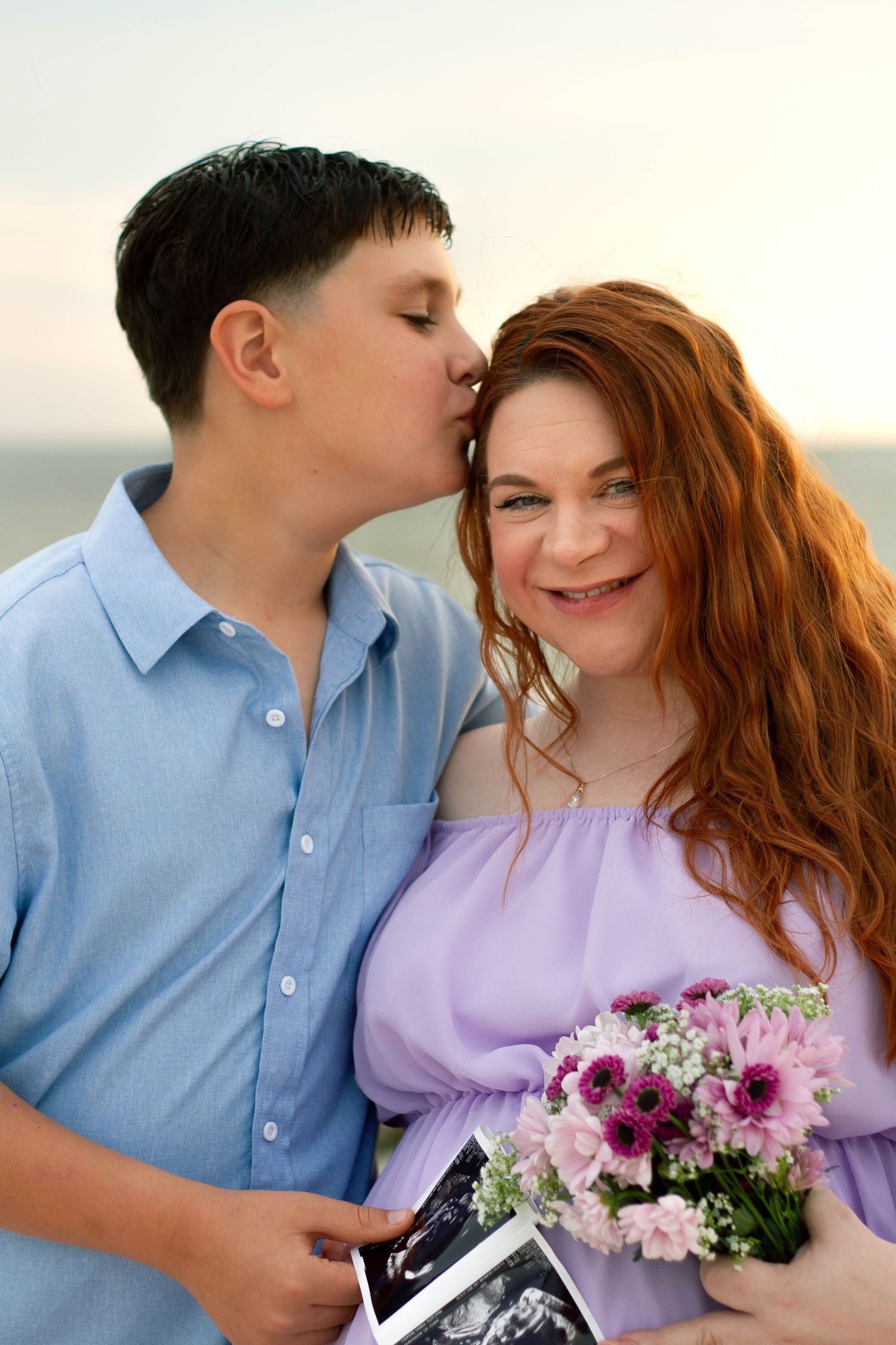 Pregnant woman in lavender dress holds flowers and ultrasound photos as her son kisses her head by the ocean.