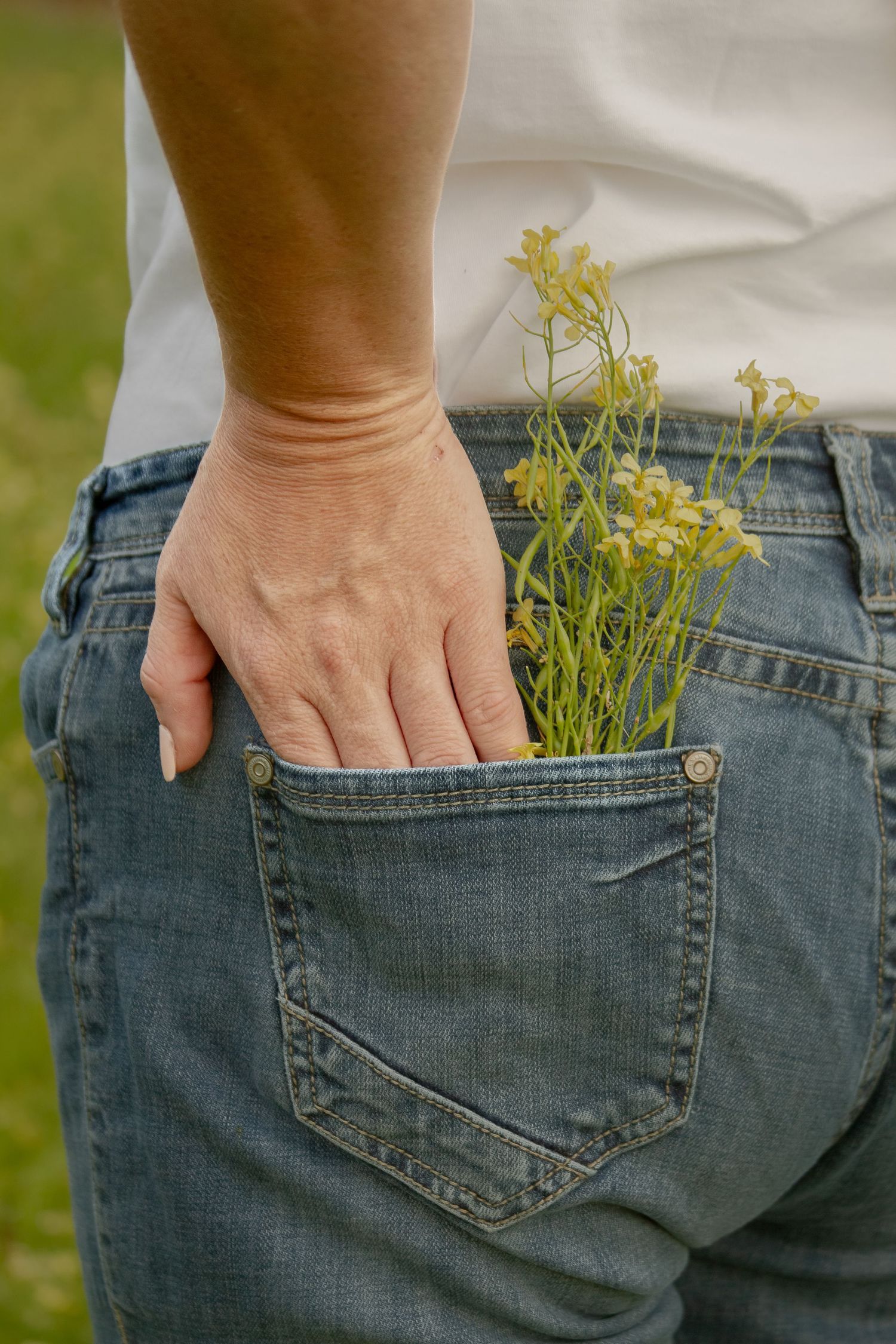 Close-up of woman in jeans with wildflowers tucked into back pocket, standing in green field.