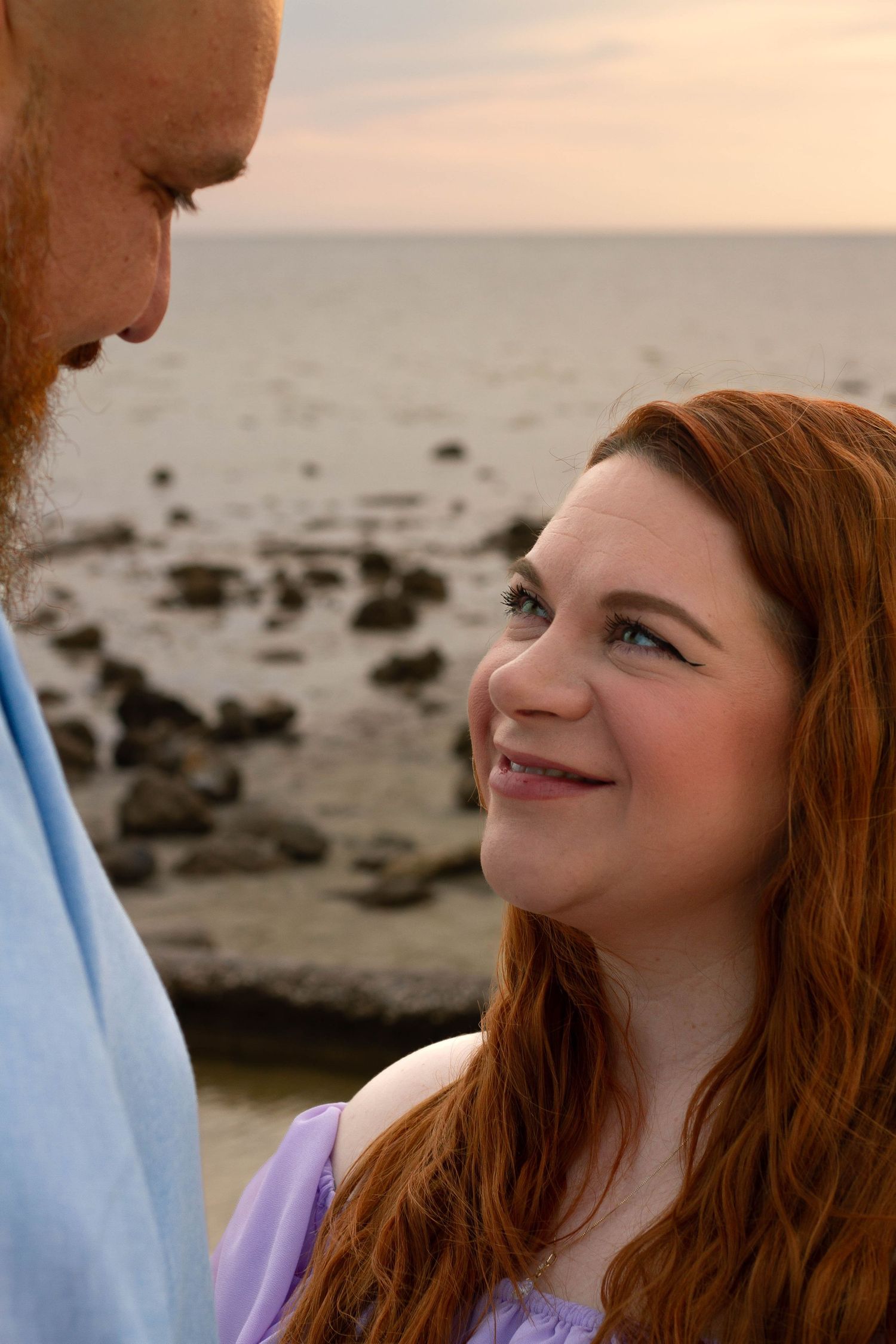 Redhead pregnant woman smiles up at bearded husband during romantic beach maternity session.