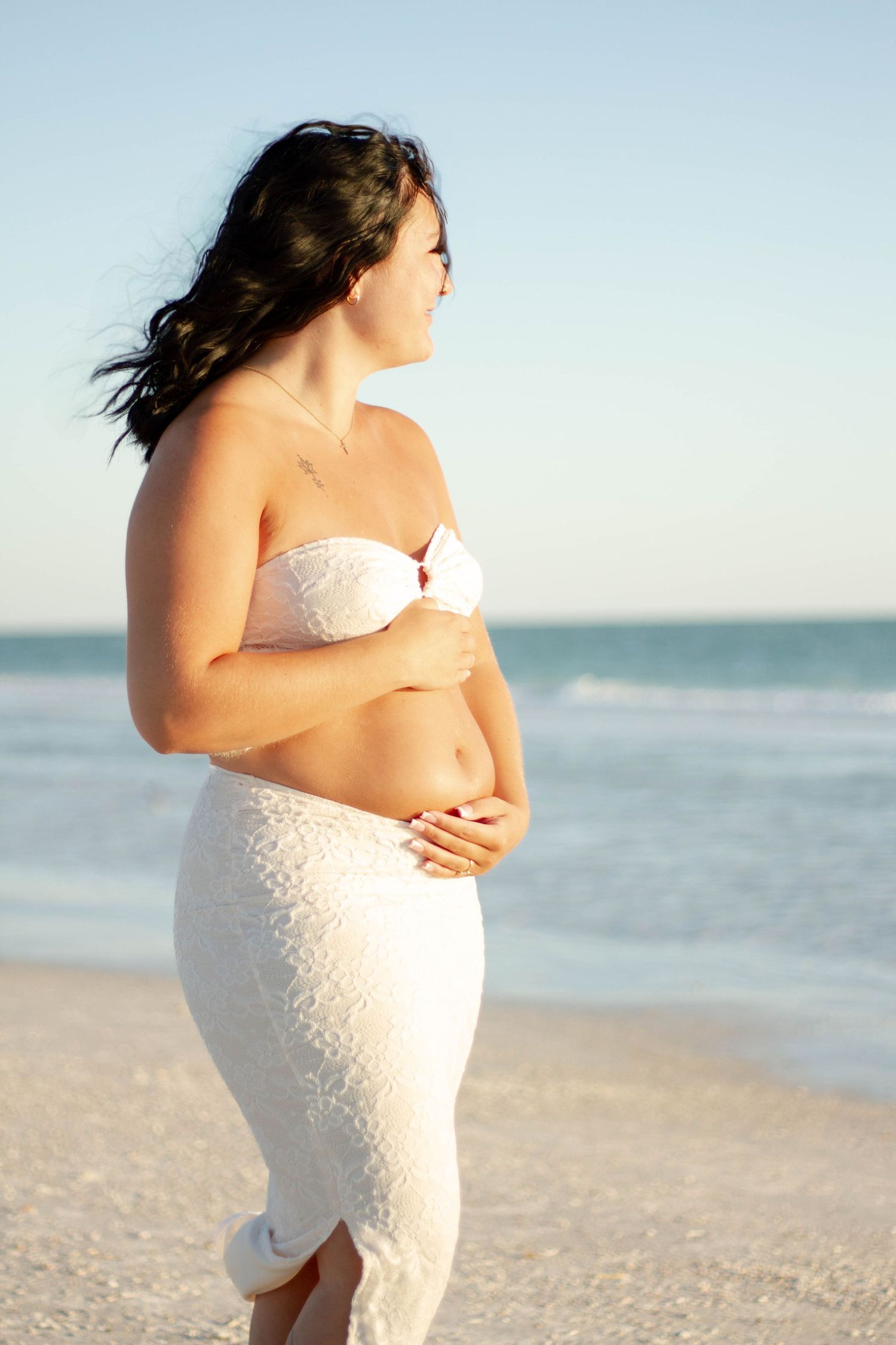 Pregnant woman in white lace skirt and bandeau top cradling baby bump on sunny beach by ocean.