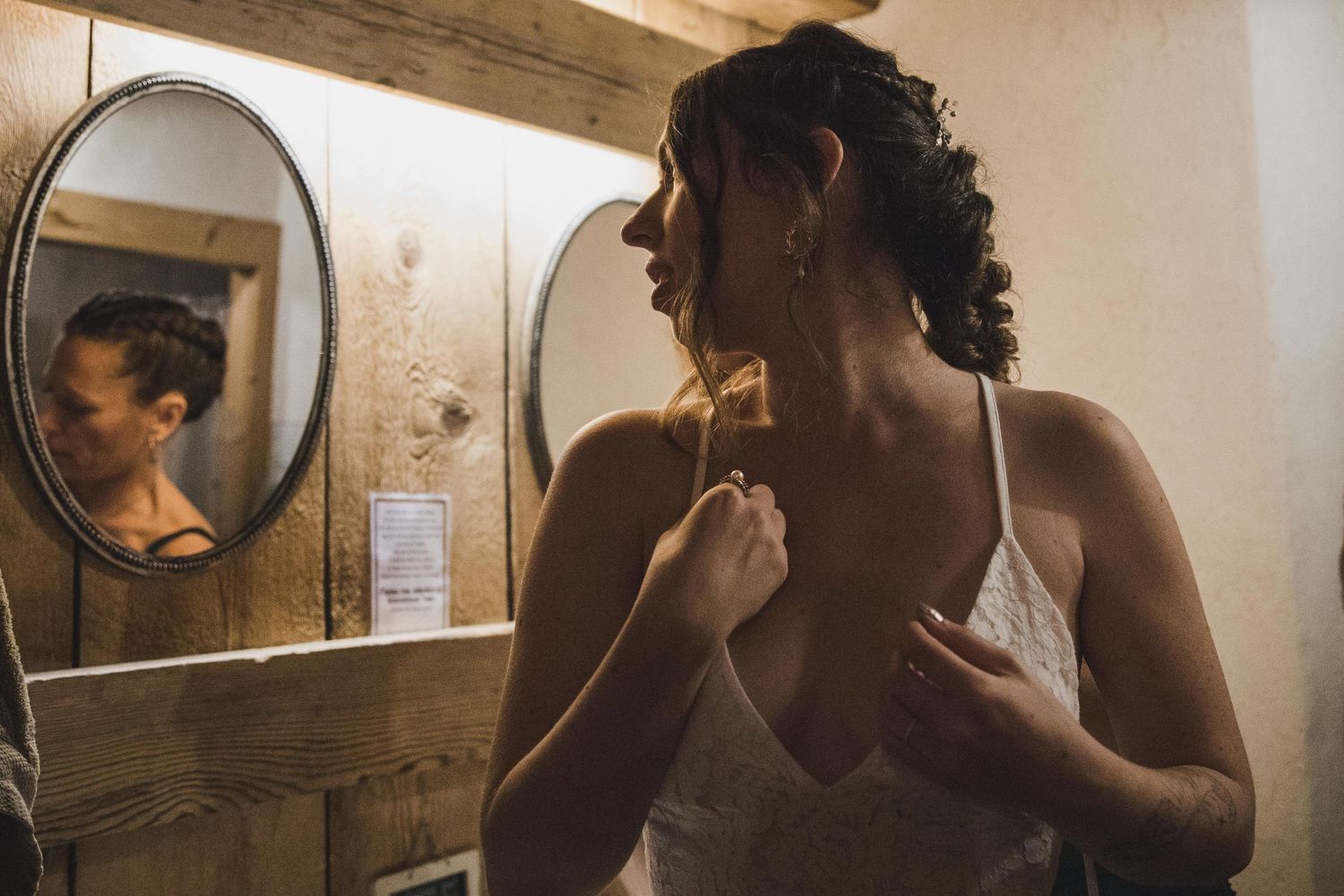 Bride adjusting earring while looking in mirror in rustic wooden bathroom.