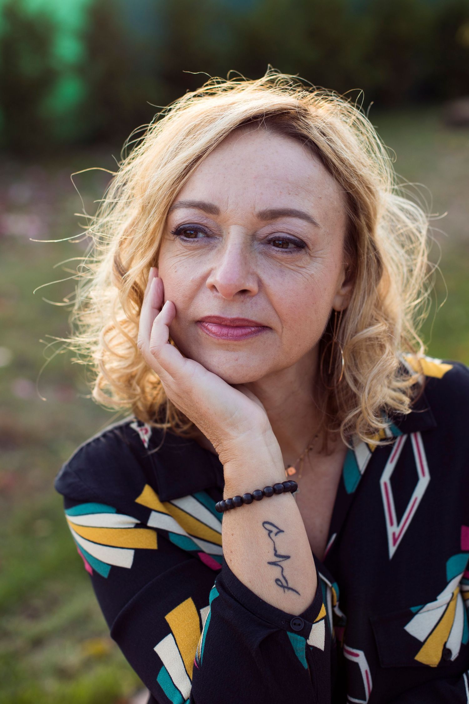 Close-up portrait of a blonde woman resting her cheek on her hand, wearing a colorful patterned blouse outdoors.