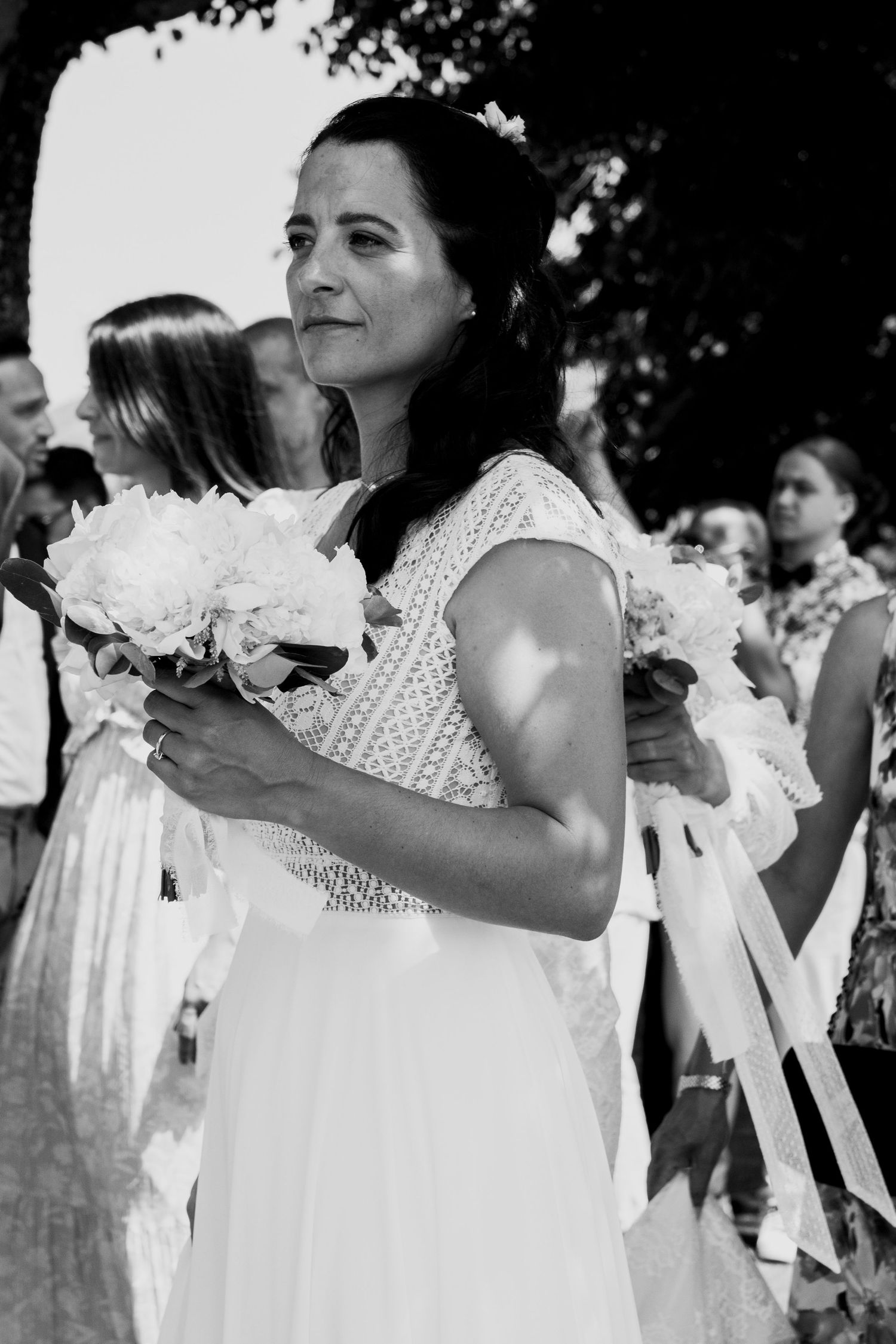 A bride in a white lace dress holds a white peony bouquet outdoors during a wedding ceremony.