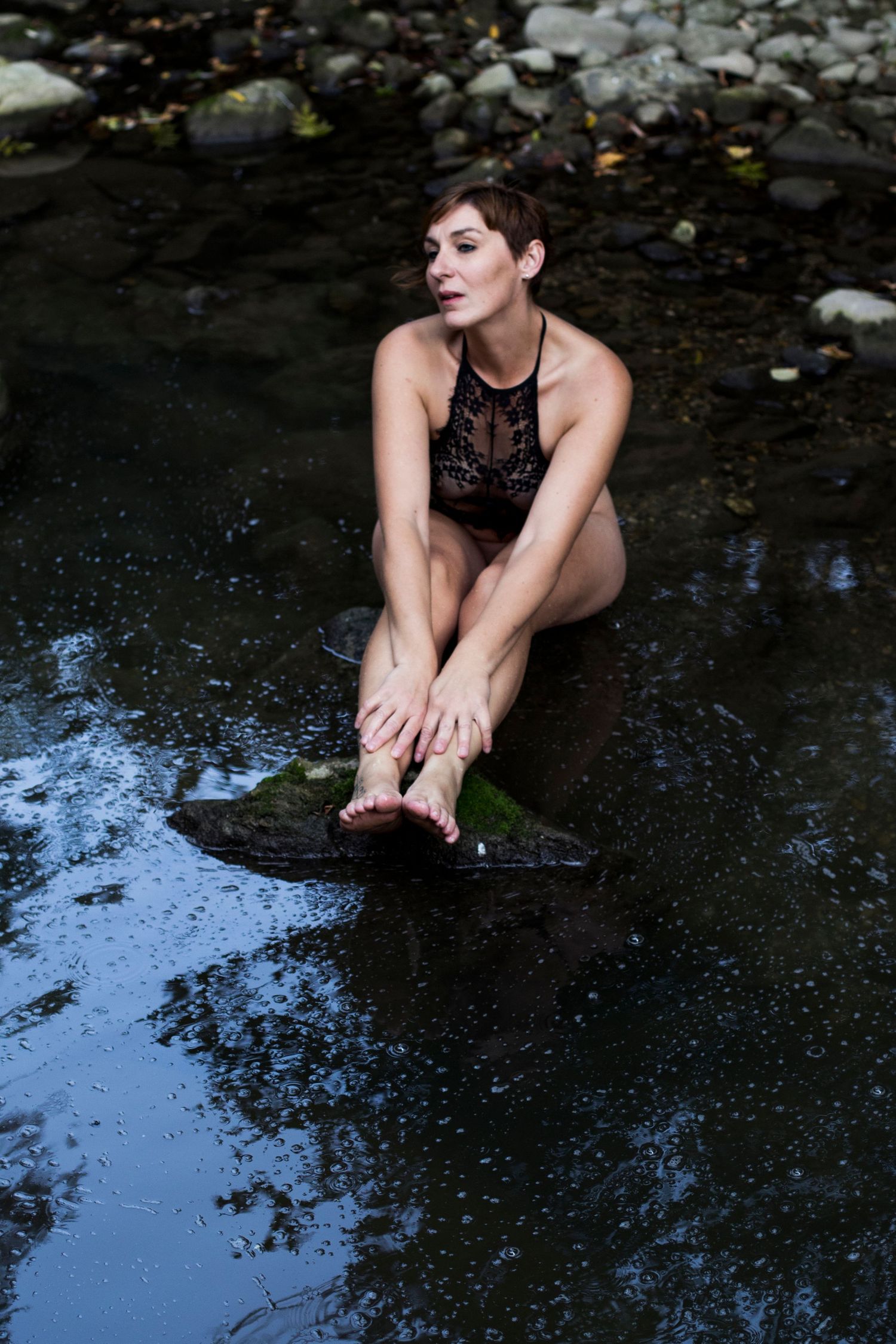 Woman in black lace bodysuit sitting in a rocky forest stream.