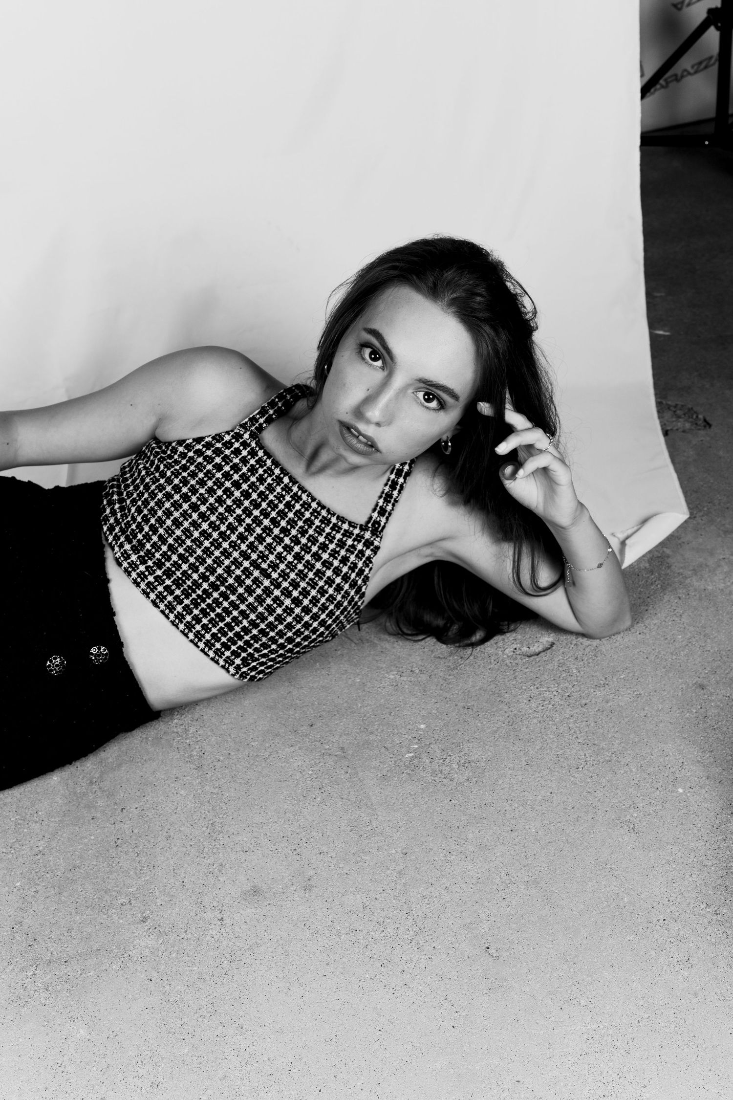 Black and white photo of young woman in checkered crop top lying on floor beside white backdrop.