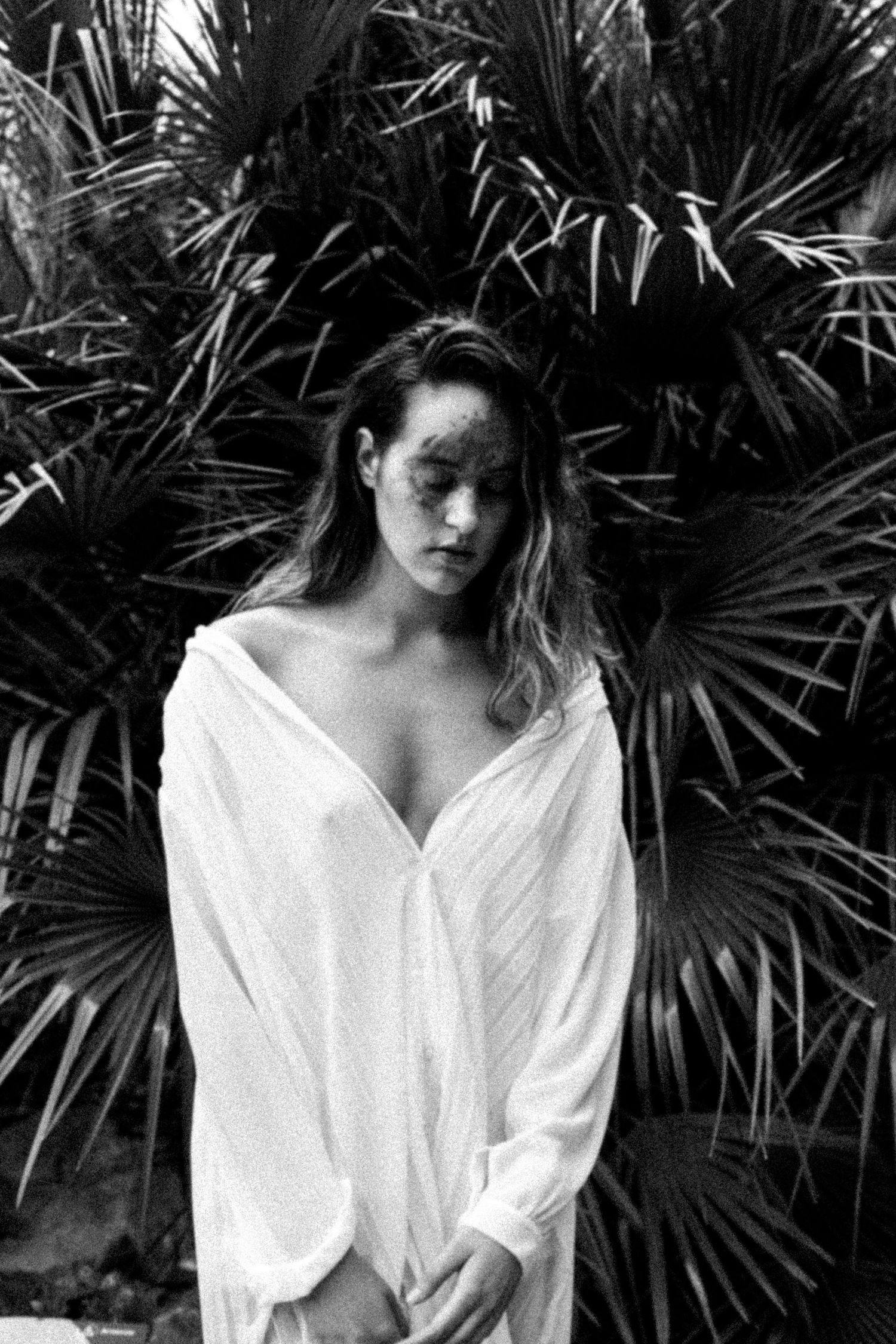 Black and white photo of a woman in an oversized white shirt standing before tropical palm leaves outdoors.