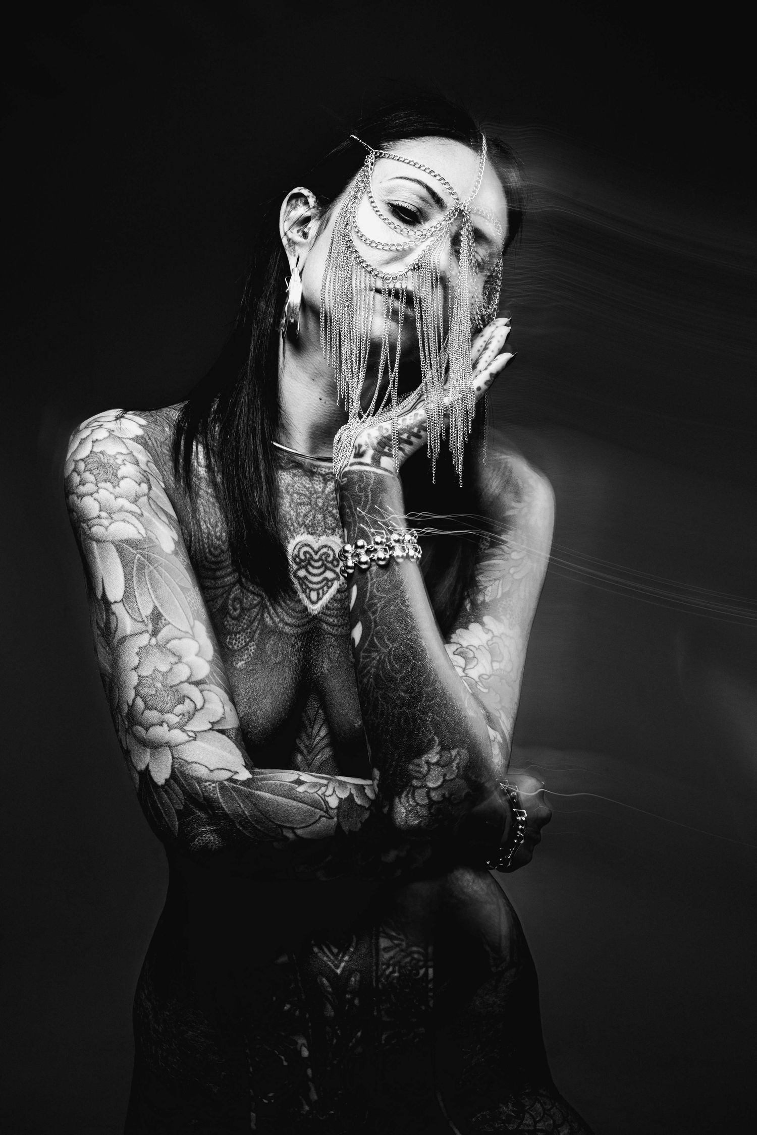 Black and white portrait of tattooed woman with gold chain face mask in moody studio light.