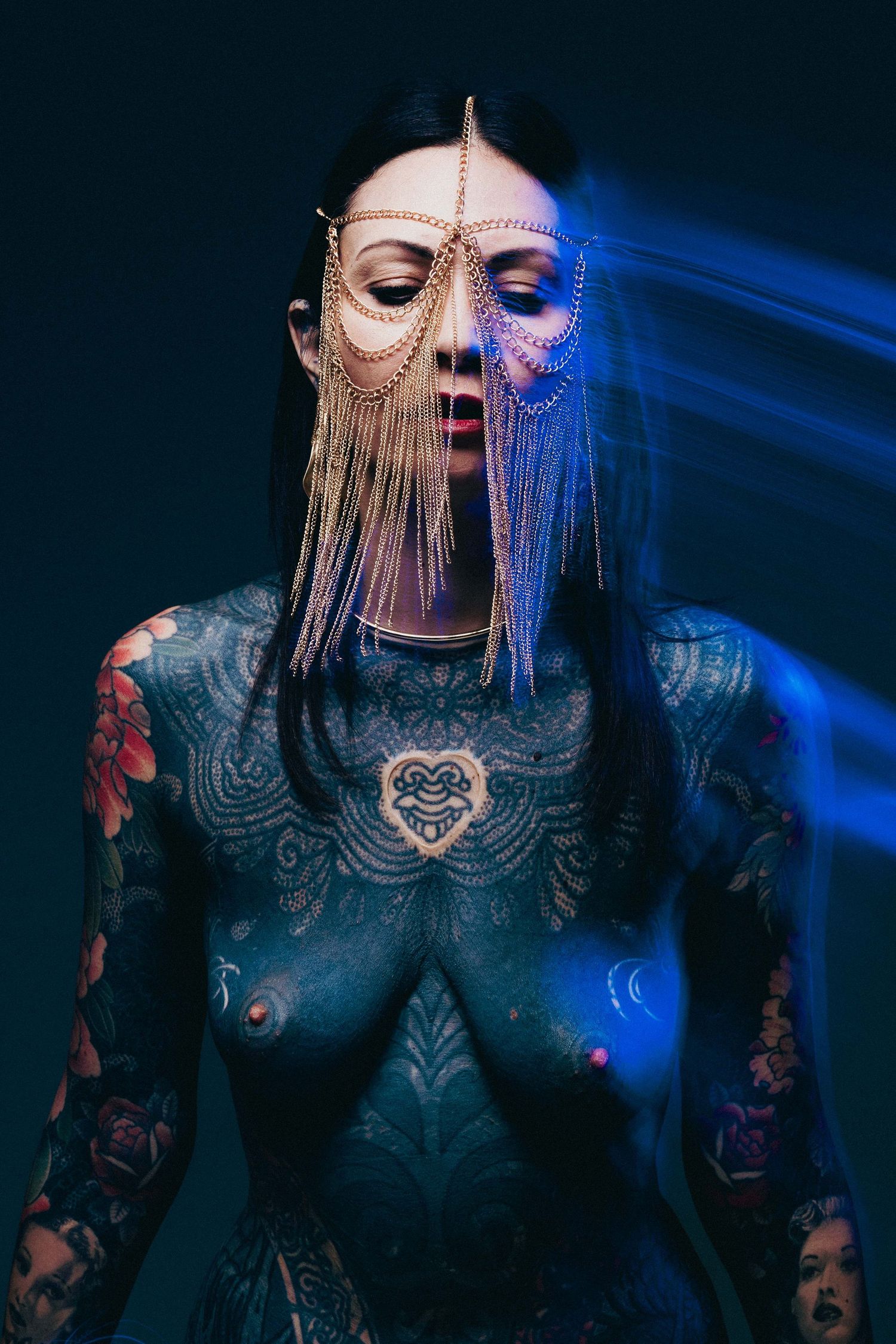 Tattooed woman wearing decorative gold chain mask with blue light streaks in dark studio setting.