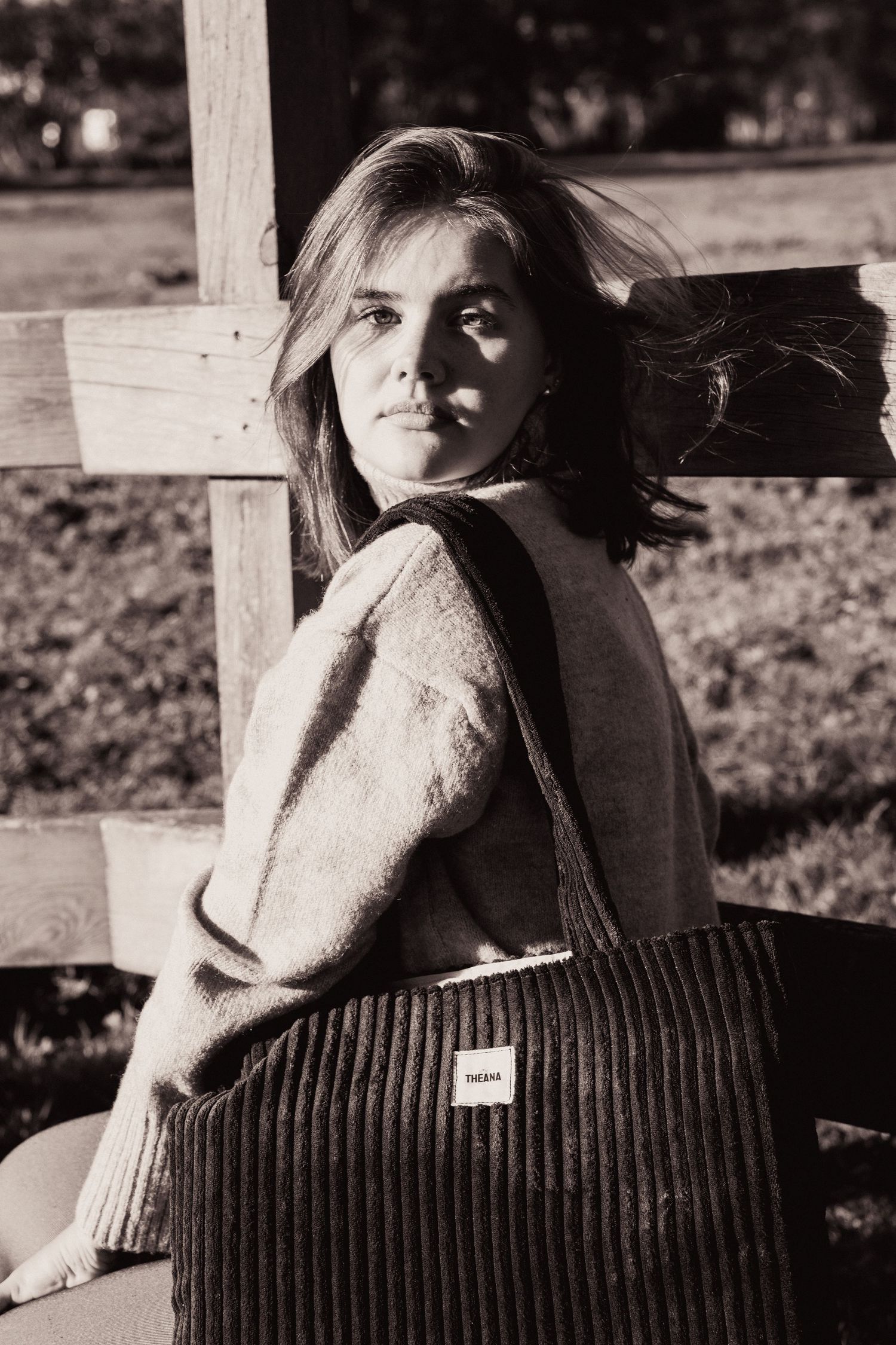 Black and white portrait of a woman in a knit sweater holding a corduroy tote bag by a wooden fence.