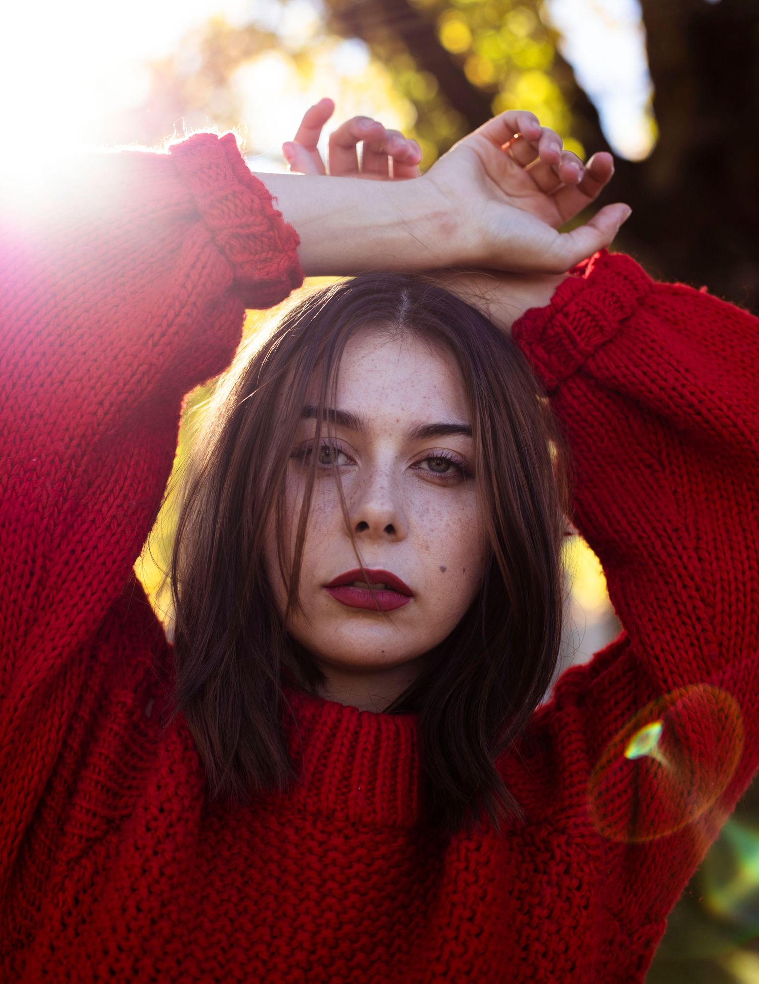 Young woman in red knit sweater poses with arms raised against golden sunlit bokeh background.