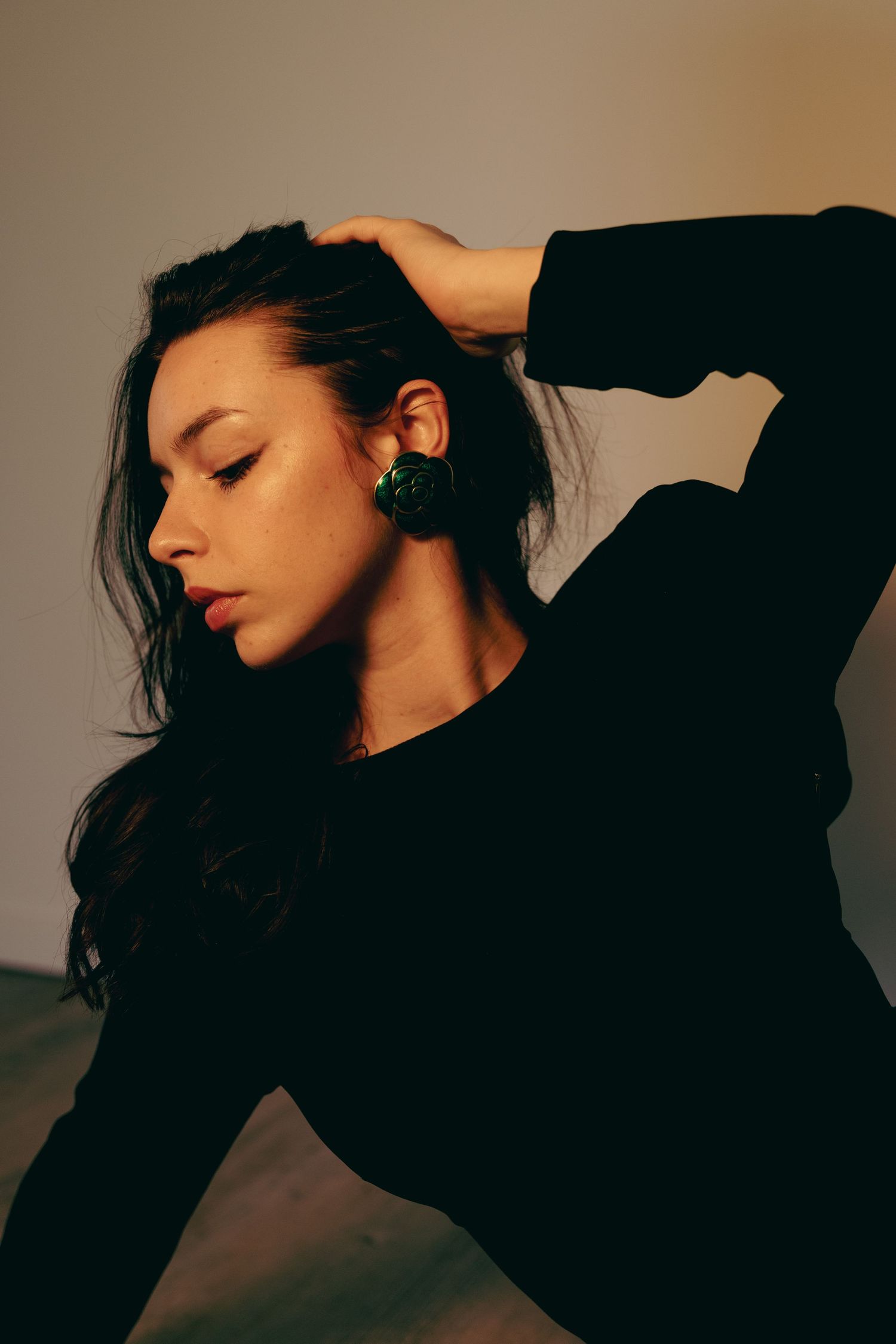 Woman in black top with green earring posing with hand in hair in warm light.