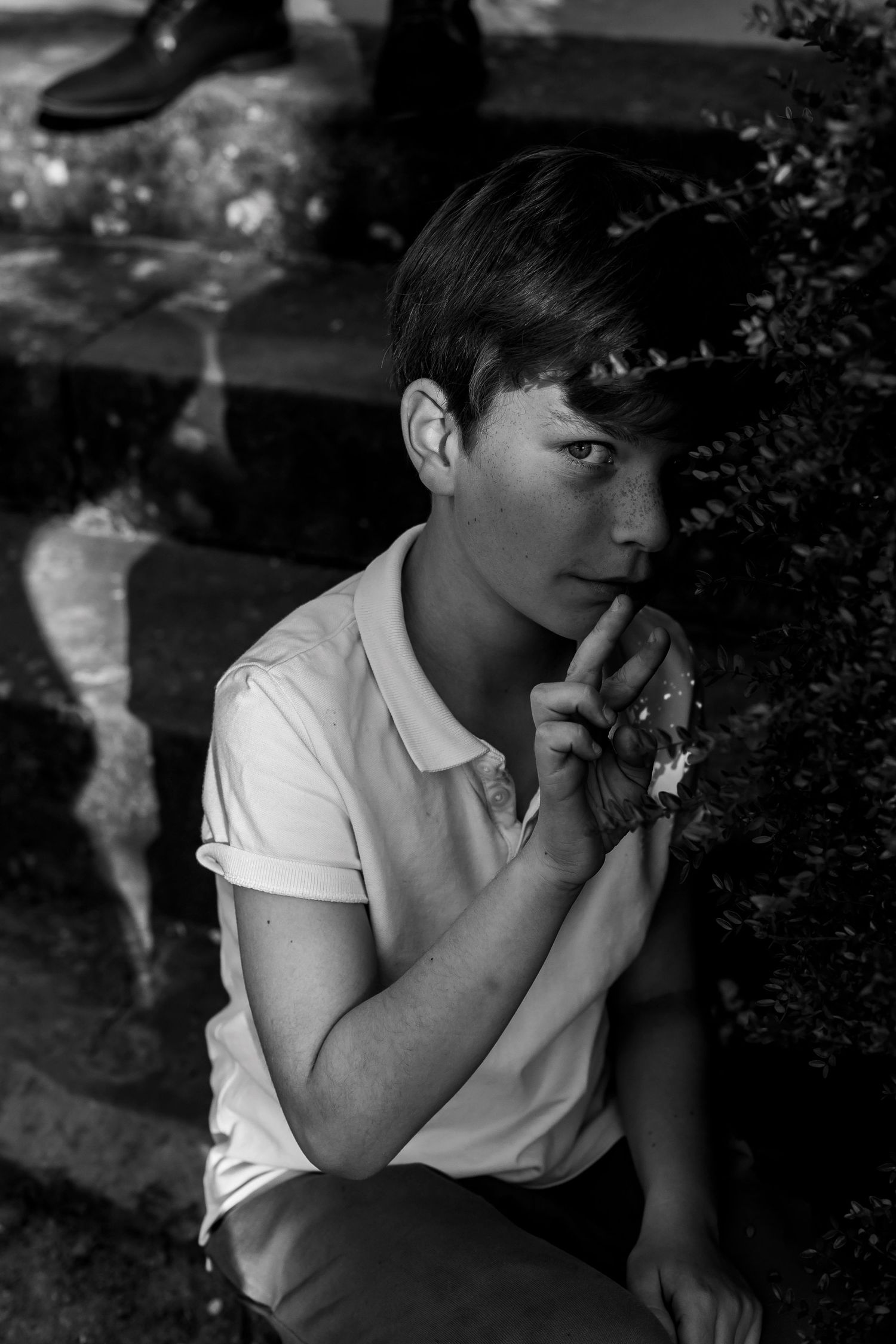 Black and white portrait of a young man sitting thoughtfully with hand near chin beside bushes.
