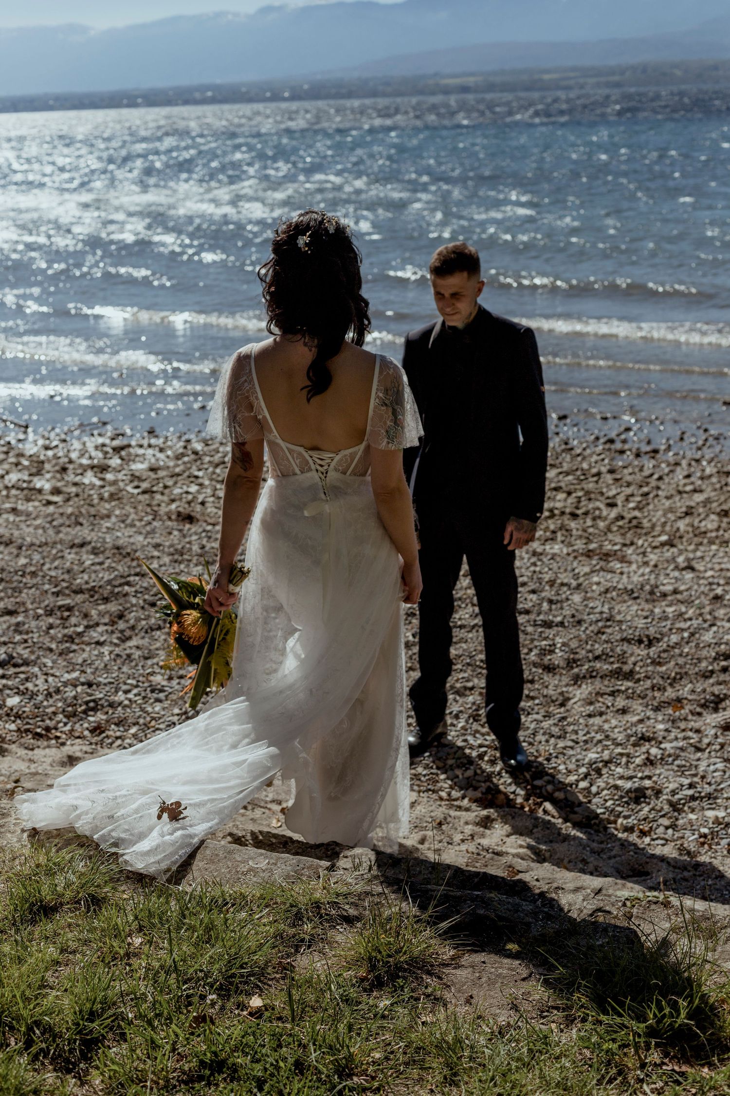 Bride in flowing white lace gown and groom in black suit pose on rocky shoreline.