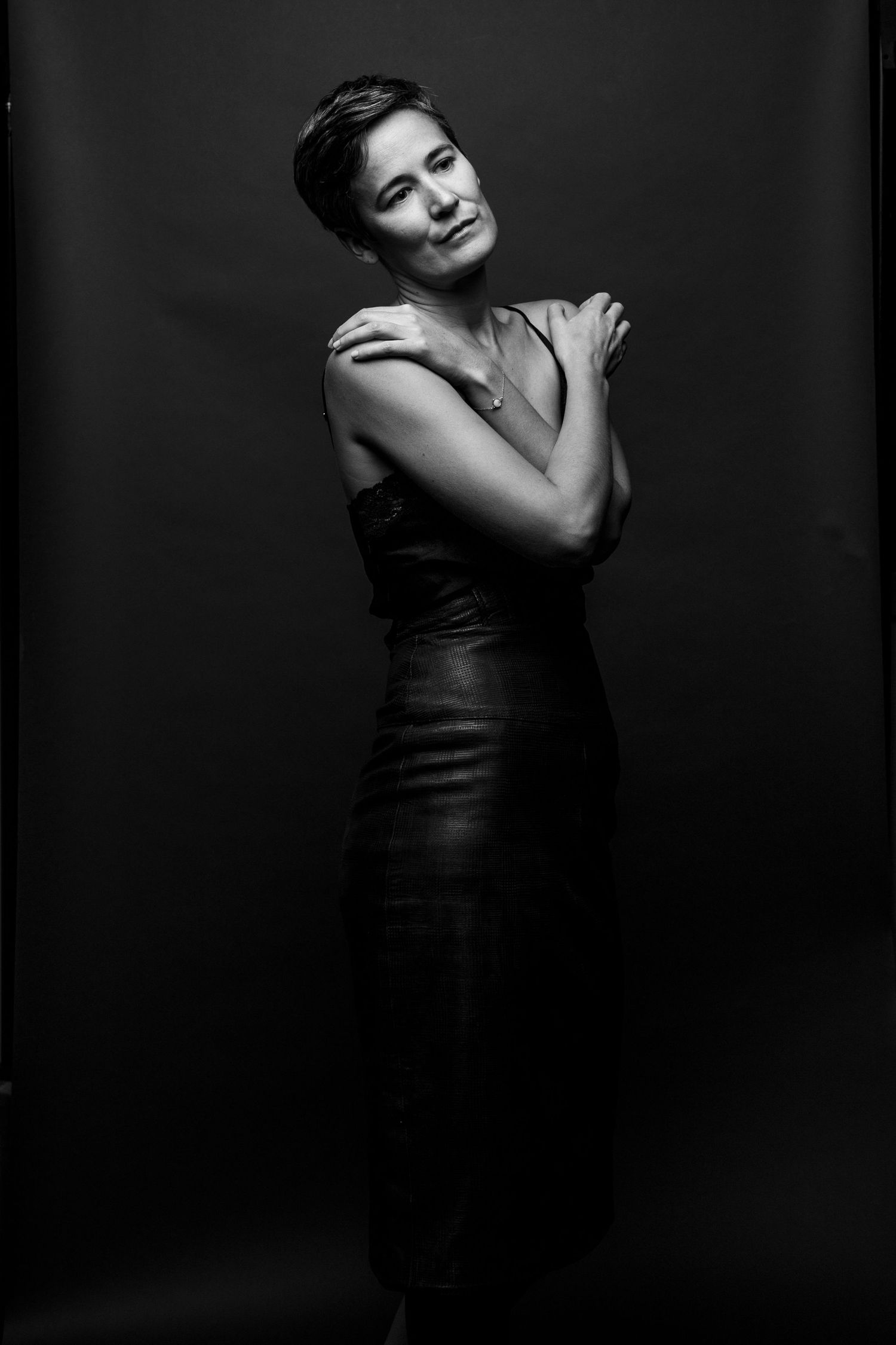 Black and white portrait of woman in sleeveless leather dress with arms crossed over chest on dark background.