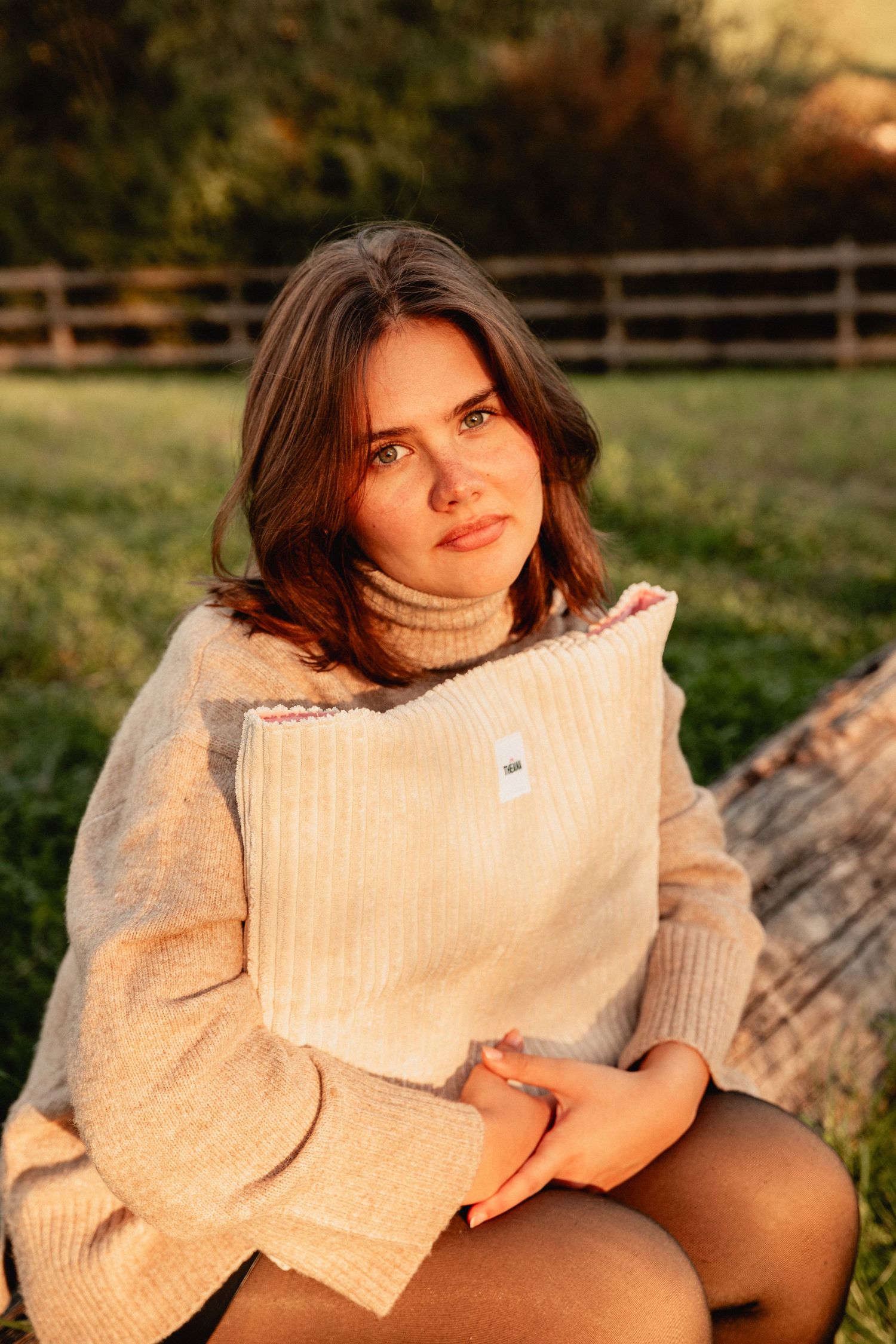 Woman in beige turtleneck sweater hugging a cream corduroy bag while sitting outdoors at sunset.