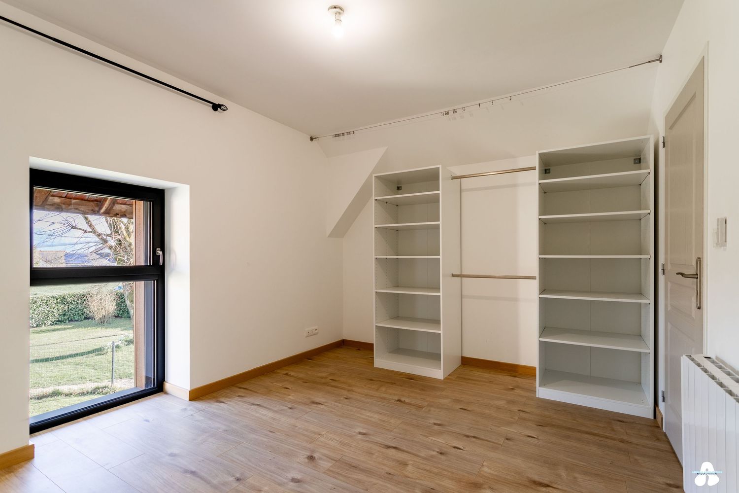 Bright attic room with built-in white shelving, wooden floor, and garden view window.