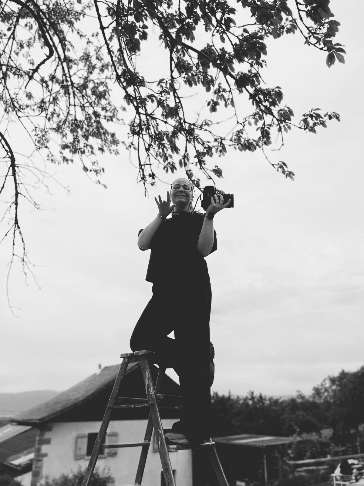 Photographer standing on ladder under tree branches, holding camera in black and white outdoor photo.