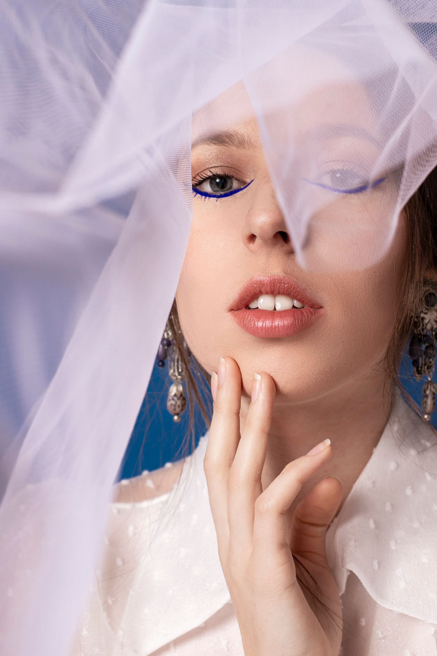 Close-up beauty portrait of a woman with blue eyeliner and jewelry partially covered by a white veil.