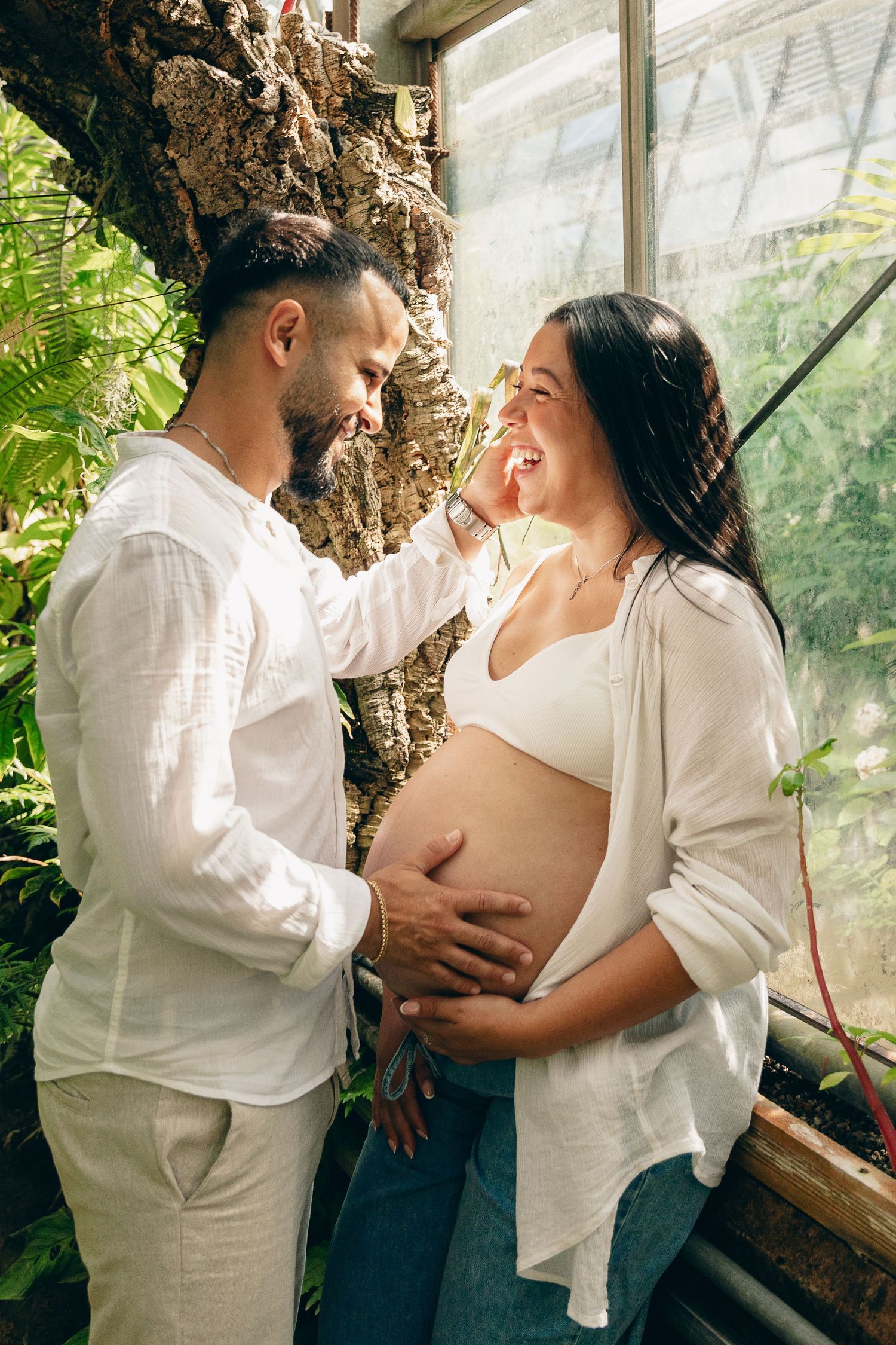 Man lovingly touches pregnant woman's face as she laughs in a sunlit greenhouse.