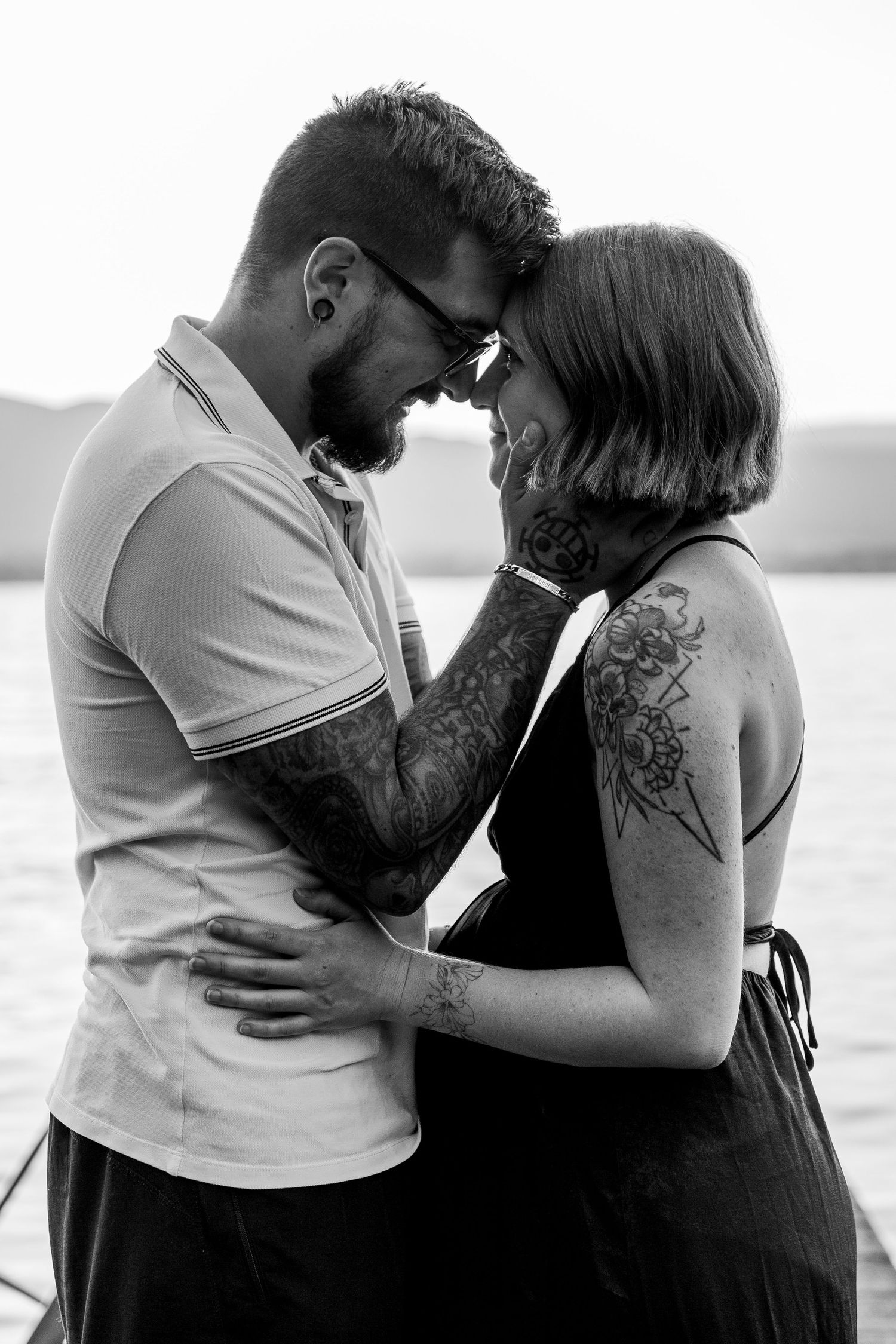 Tattooed couple sharing intimate moment, foreheads touching near lake in black and white photo.
